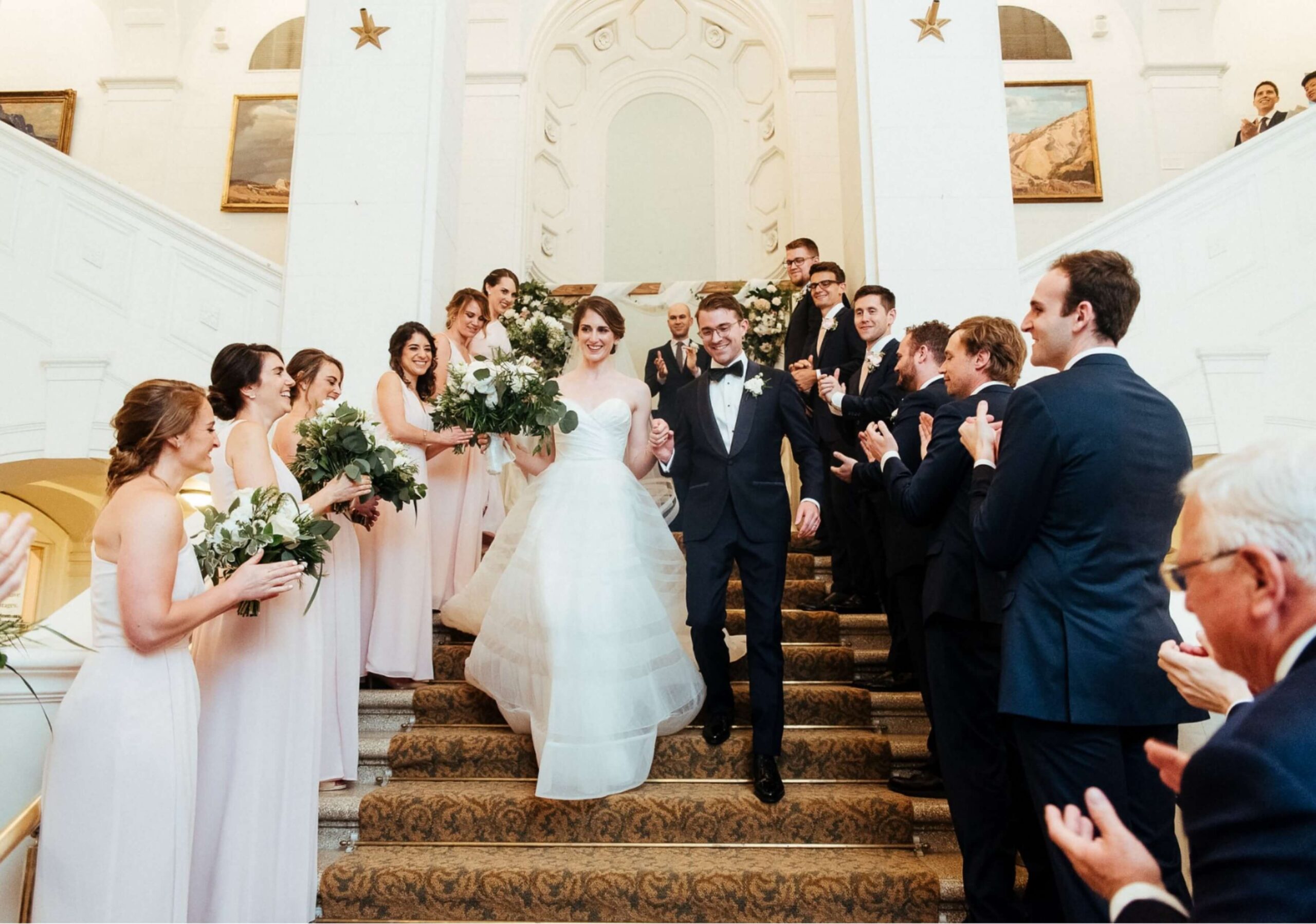 A bride and groom descend a grand staircase surrounded by bridesmaids and groomsmen holding bouquets, amid applause in an elegant venue.