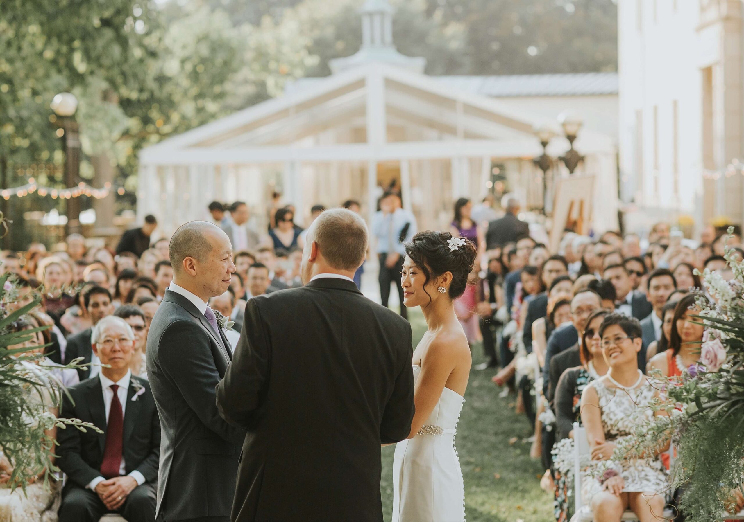 A couple in wedding attire stands before an officiant during an outdoor ceremony, with guests seated on either side.