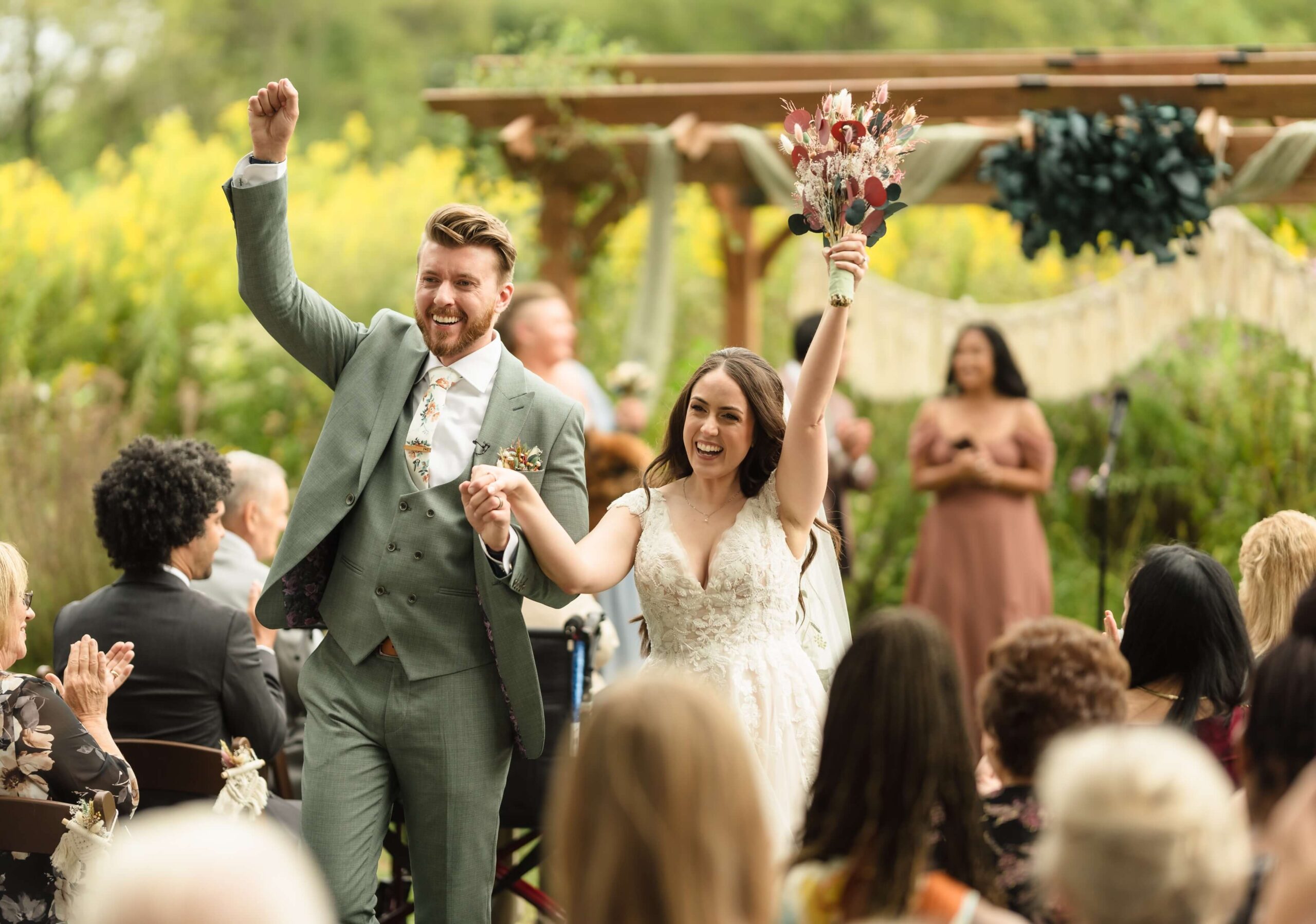 A couple walks down the aisle outdoors, cheering and holding hands. The bride holds a bouquet; guests are seated. Decor includes flowers and a wooden arch.