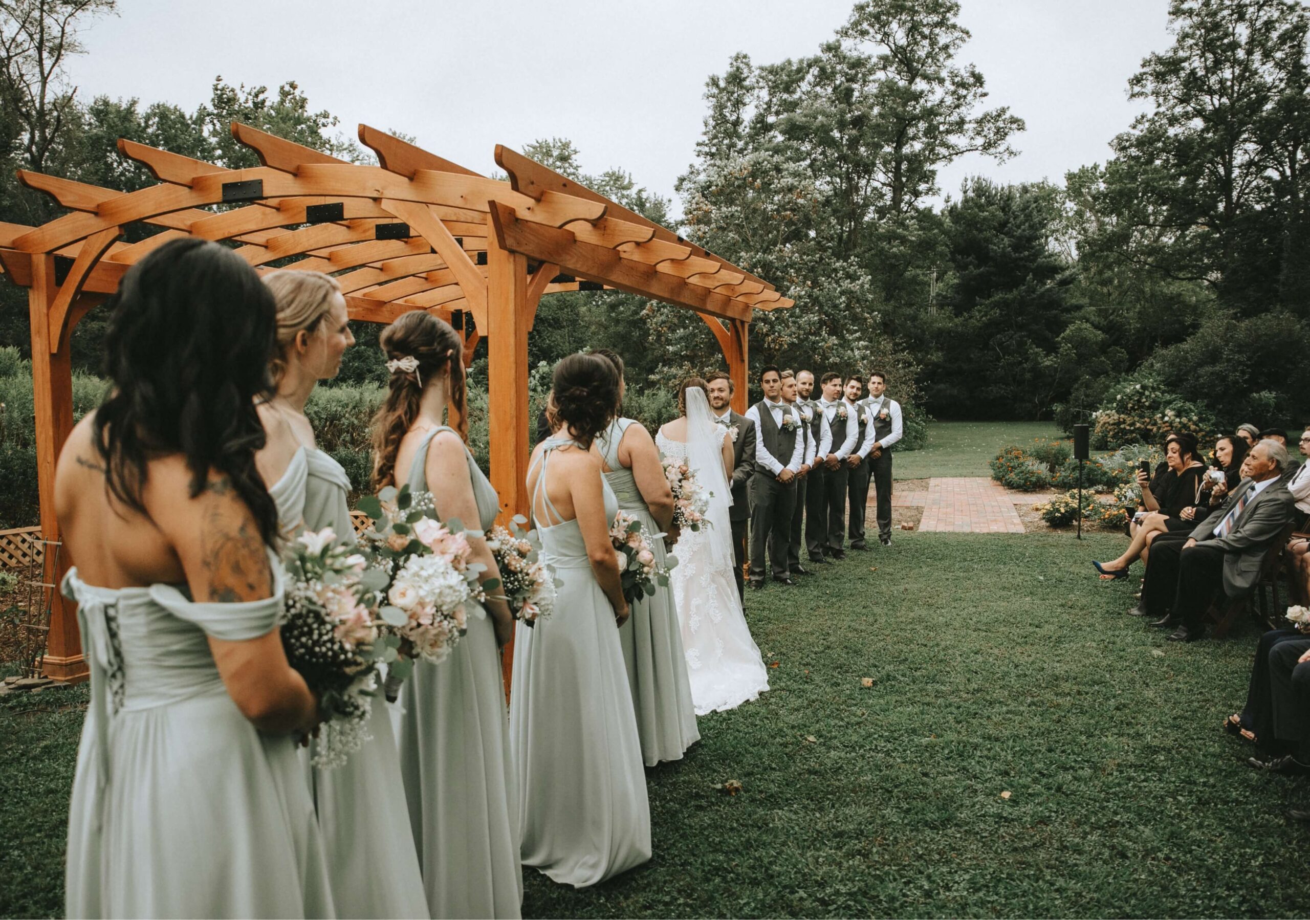A wedding ceremony outdoors with bridesmaids in light dresses and groomsmen in vests. The couple stands in front of a wooden arch, and guests are seated on the right side.
