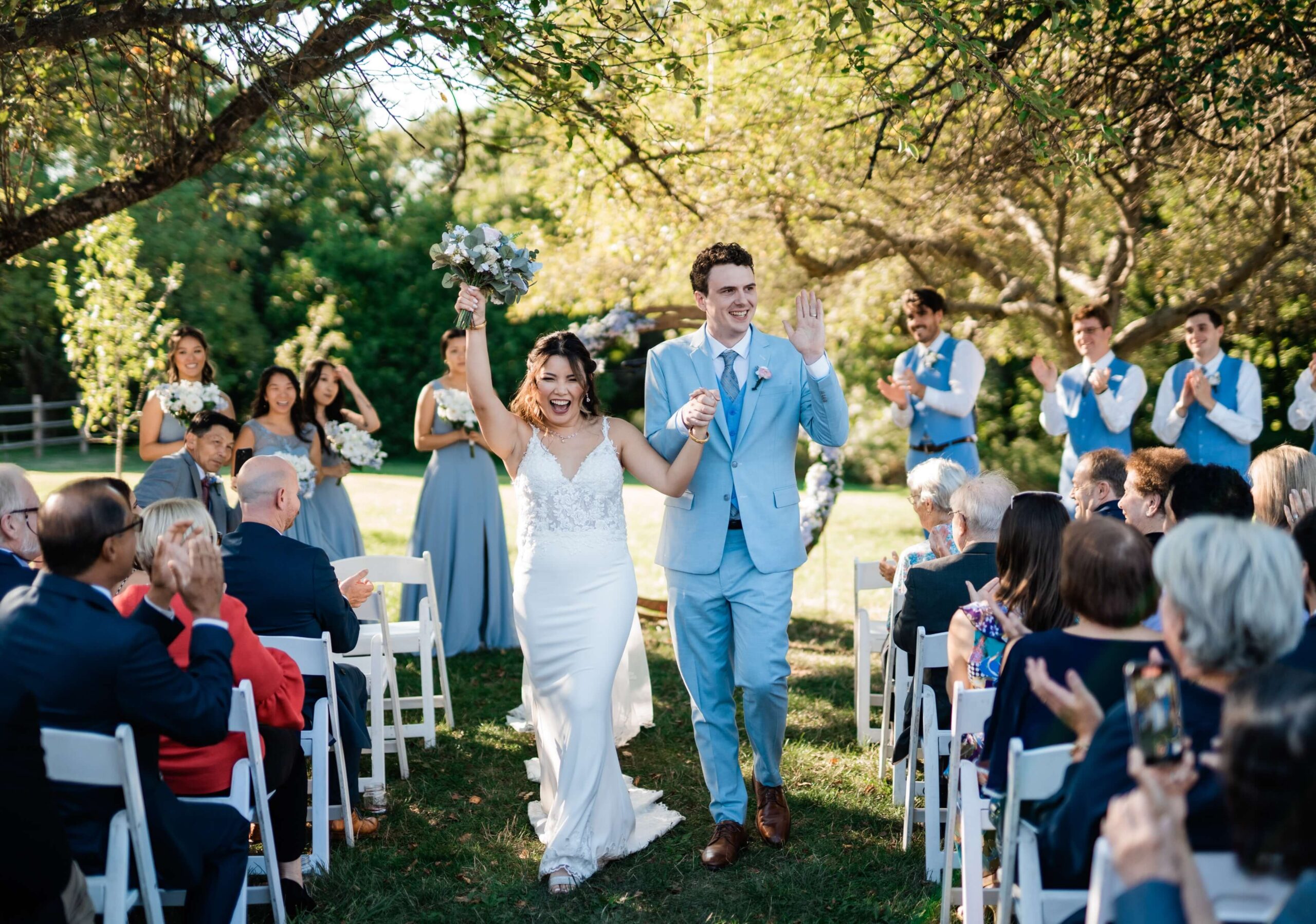 Bride and groom walk down the aisle, smiling and celebrating, with guests seated on either side. Bridesmaids and groomsmen stand clapping in the background under sunlit trees.