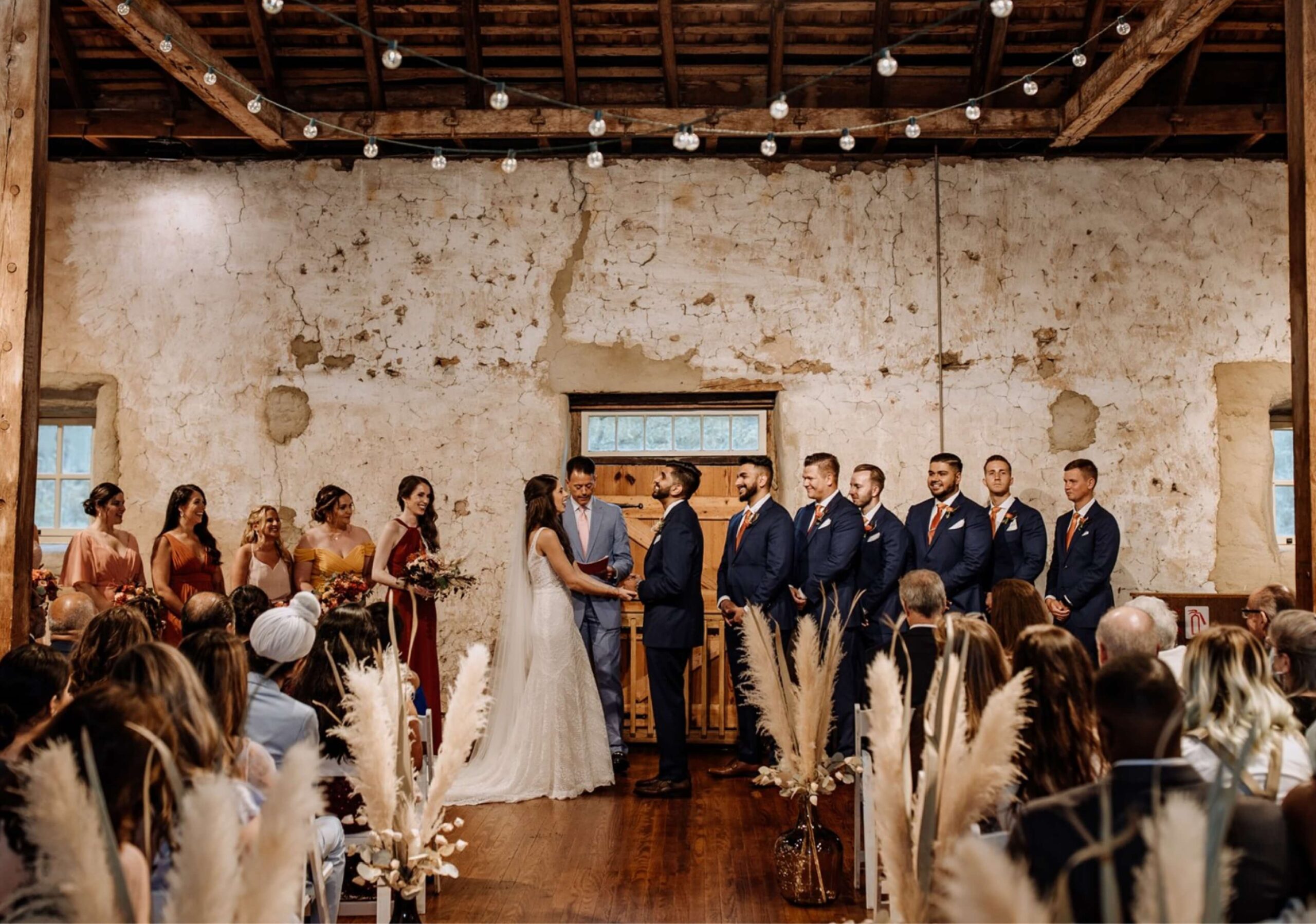 A wedding ceremony in a rustic venue with exposed beams. A couple stands at the altar surrounded by their bridal party, while guests are seated, viewing the proceedings.