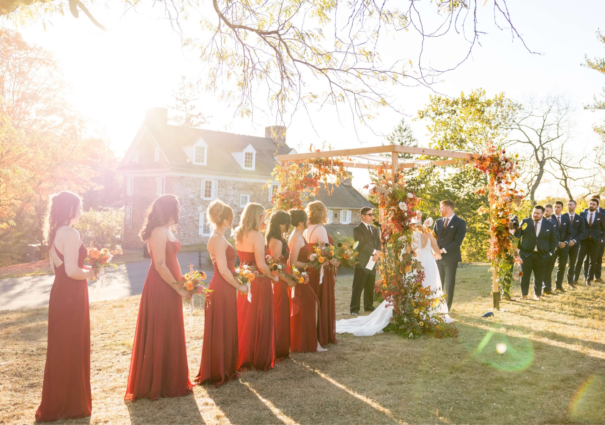Wedding ceremony outdoors with bridesmaids in red dresses and groomsmen in suits. The couple stands under a floral arch, and guests are seated under a sunny sky.