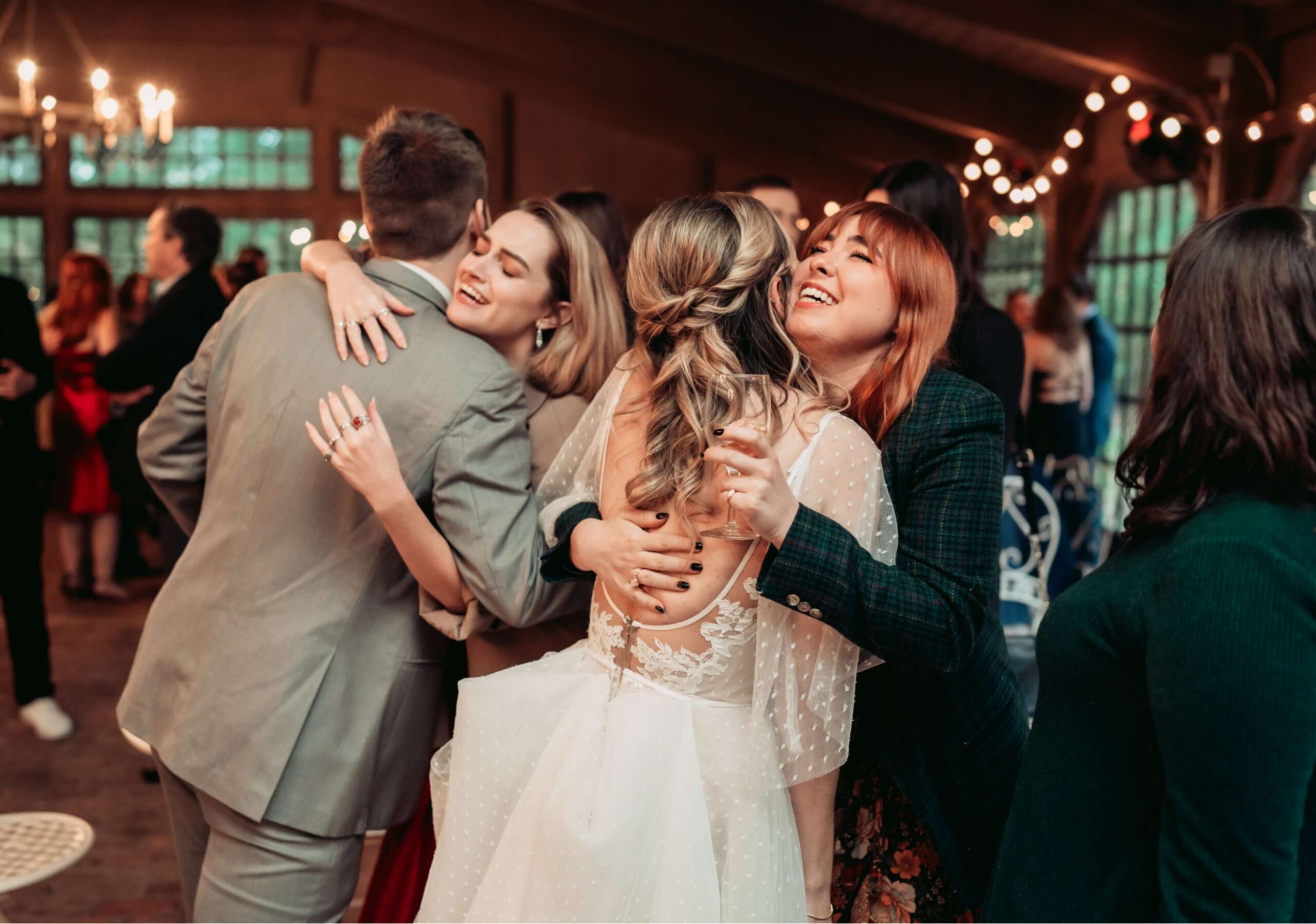 A group of people, including a couple in wedding attire, joyfully hug each other in a warmly lit venue.