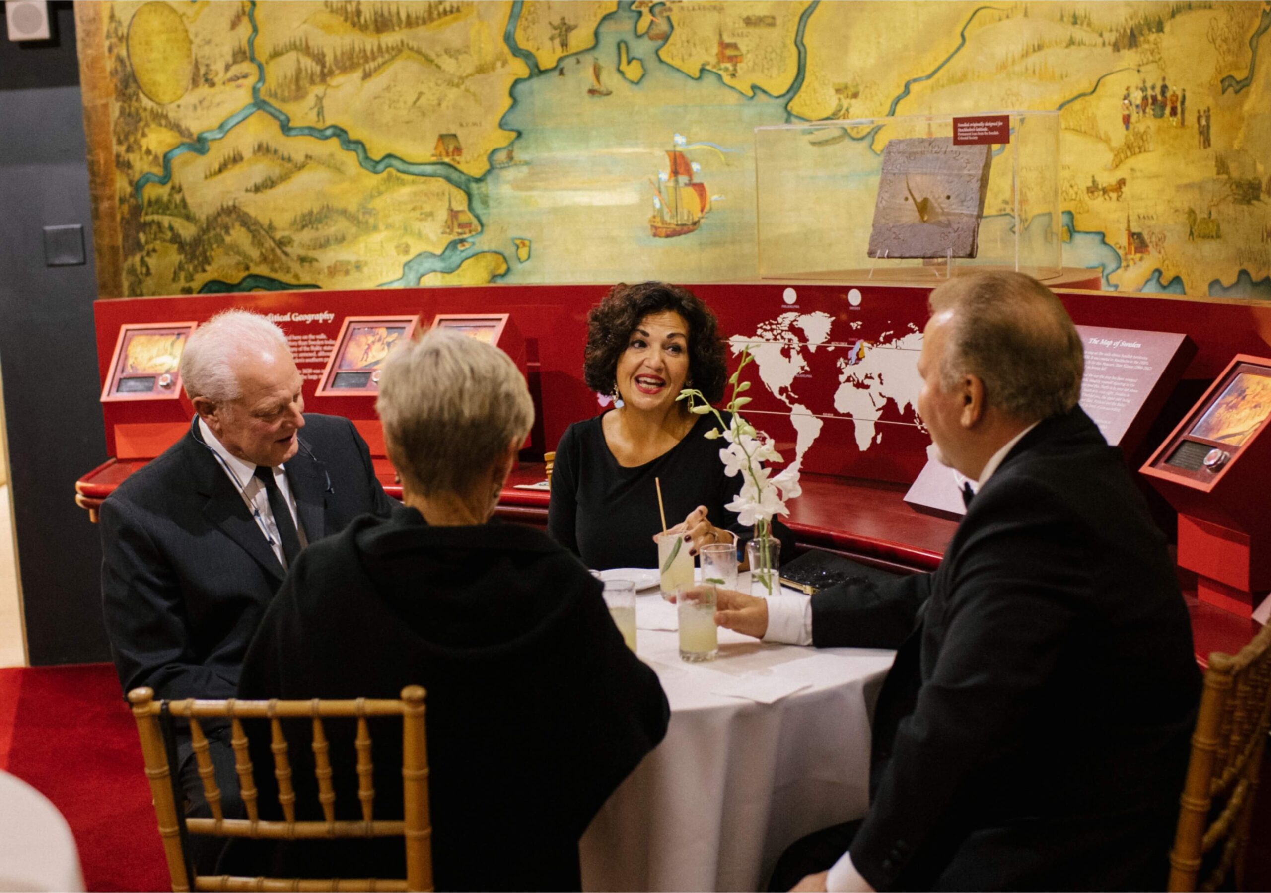 Four people sit around a table, conversing and smiling, in a room with historical maps on the walls and display cases nearby.
