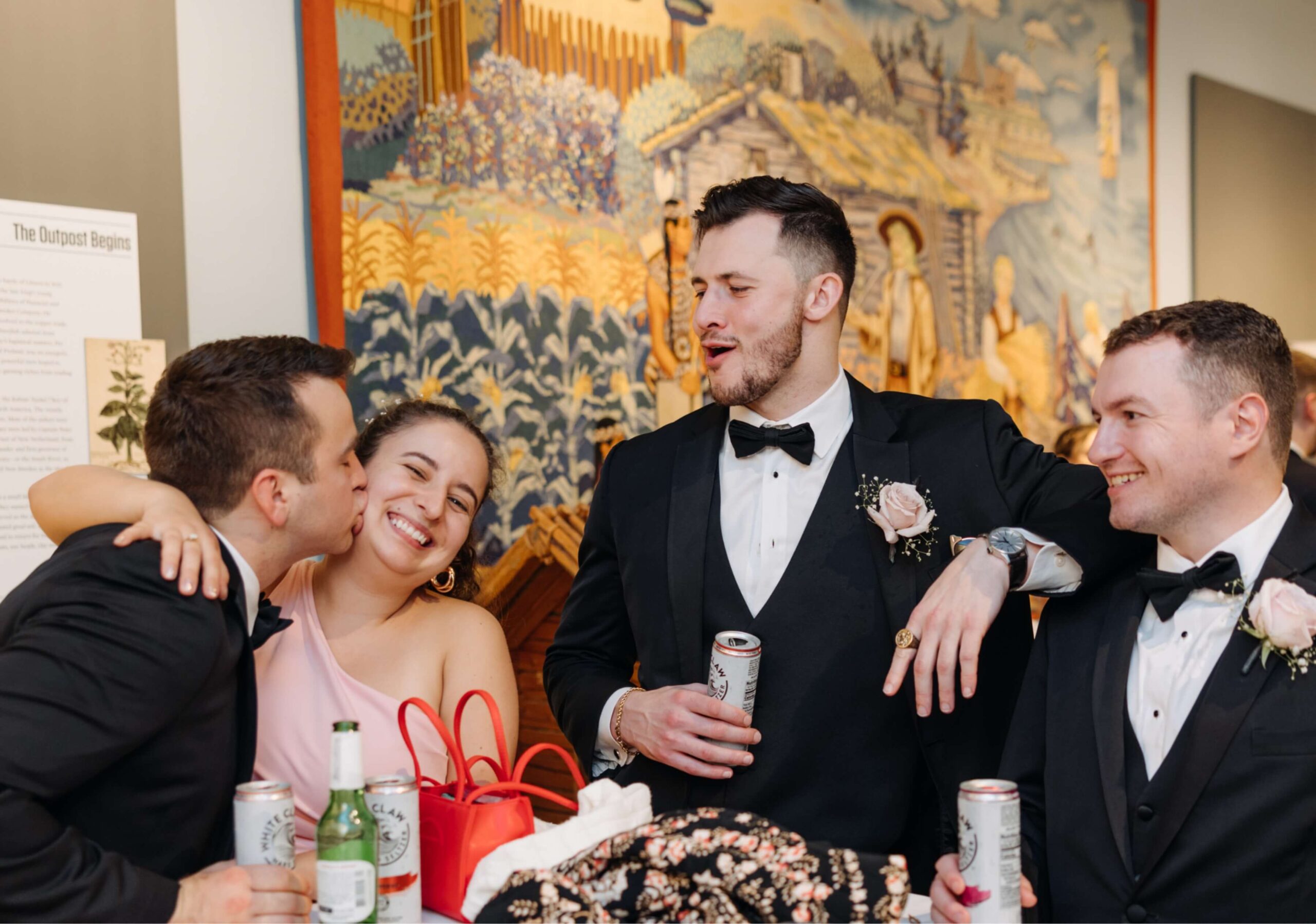 Three men and one woman in formal attire, smiling and holding drinks at an indoor gathering with a tapestry background. One man kisses the woman's cheek.