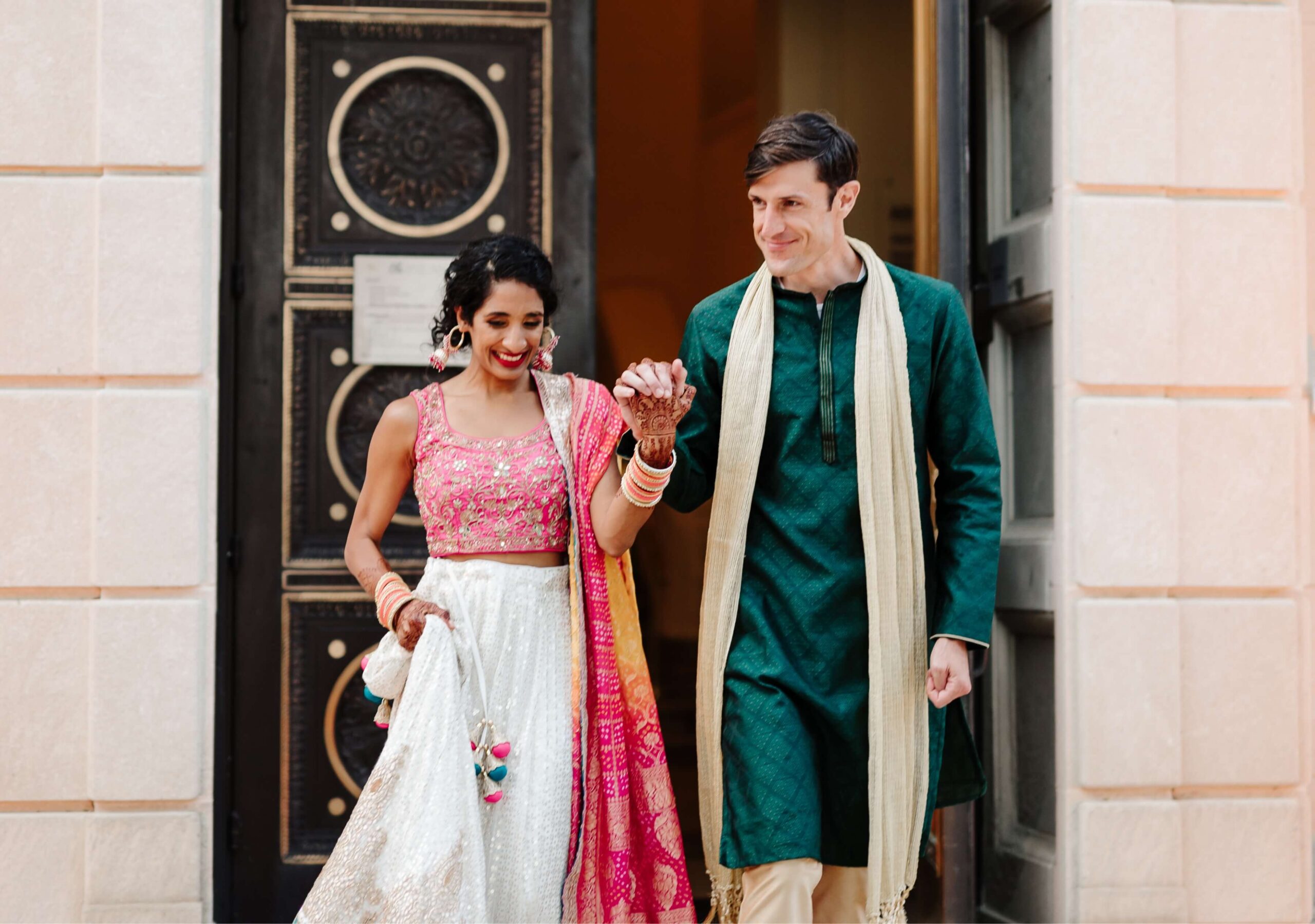 A couple in traditional attire holding hands and smiling as they walk through a doorway.