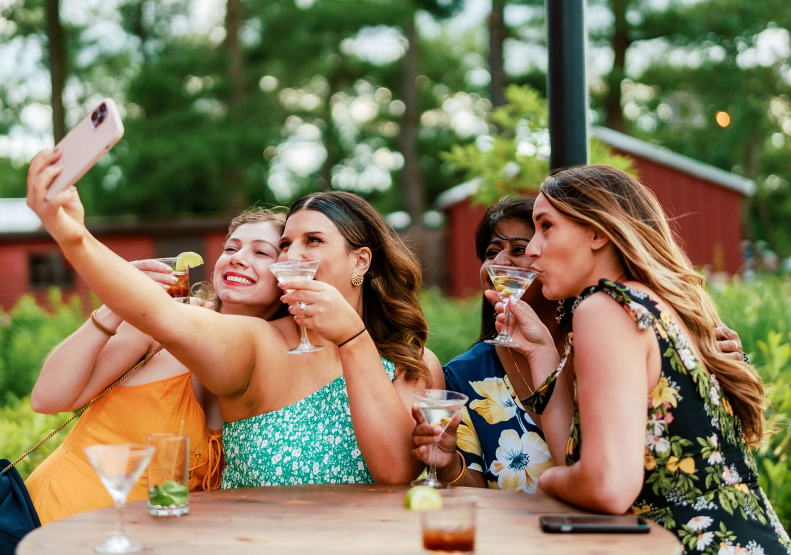 Four women sit at an outdoor table, holding drinks and posing for a group selfie. They are surrounded by greenery with blurred trees and a red building in the background.