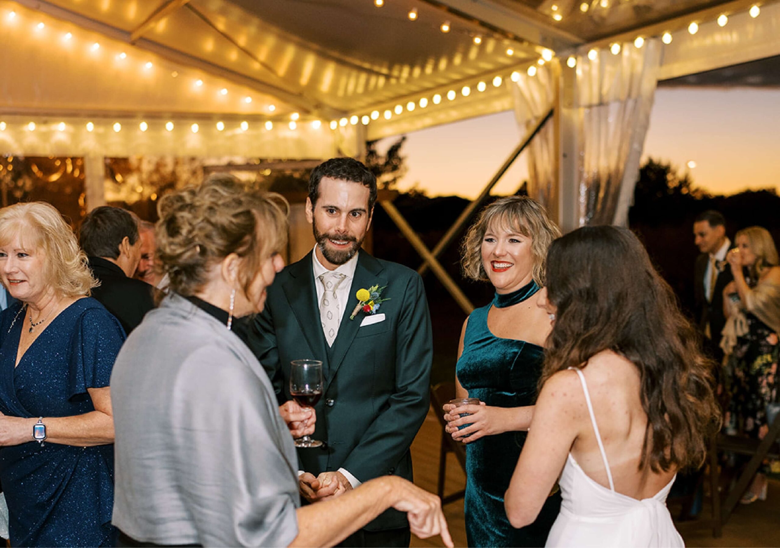 People gathered at an evening event under string lights, engaged in conversation. A man in a suit holds a glass, with women around him in formal attire.