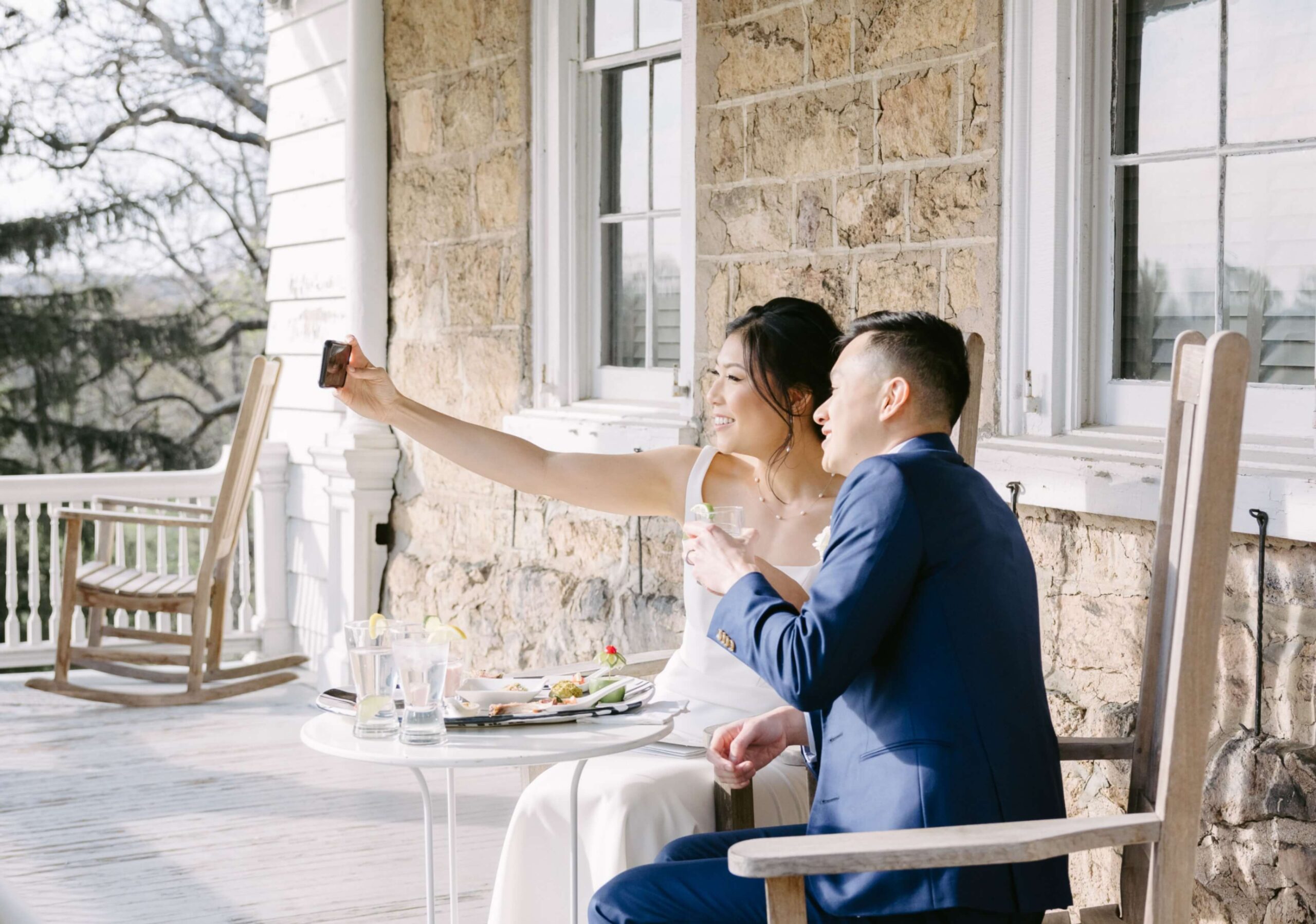 A couple in formal attire takes a selfie while sitting at a small table on a porch.