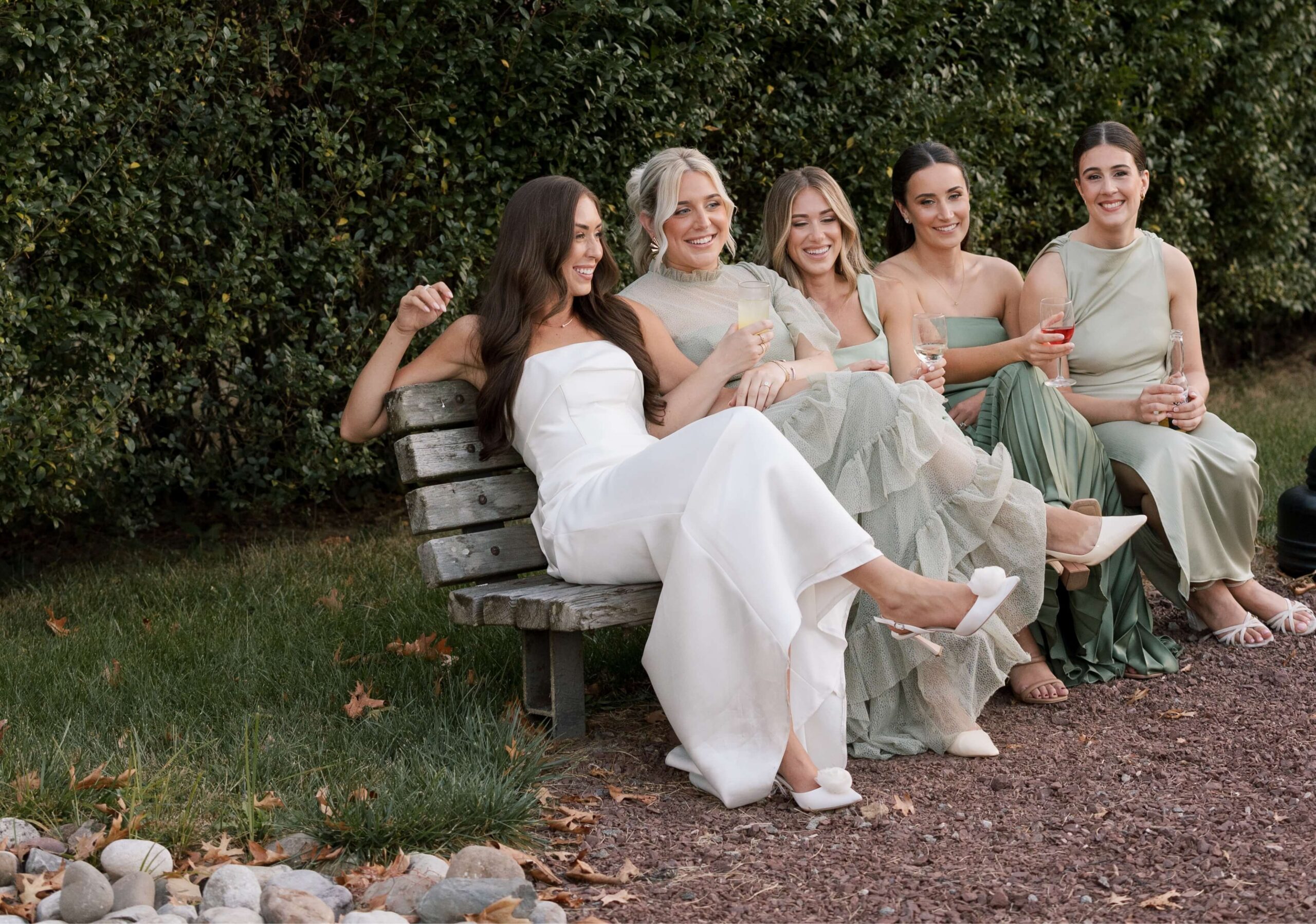 Five women in formal dresses sit on a wooden bench outdoors, holding drinks and smiling, with greenery in the background.