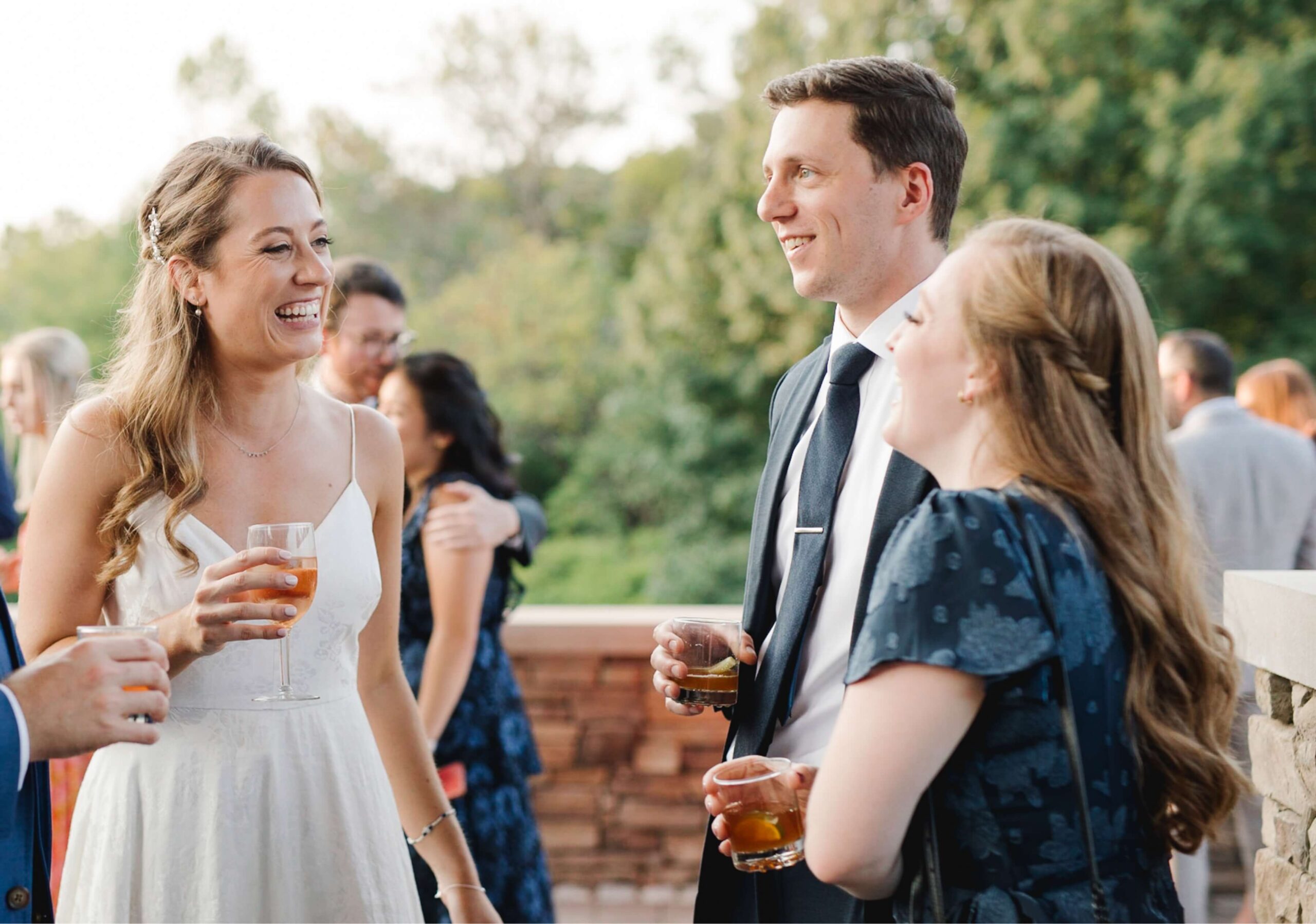 A group of people in formal attire are smiling and holding drinks while standing outdoors, with trees in the background.