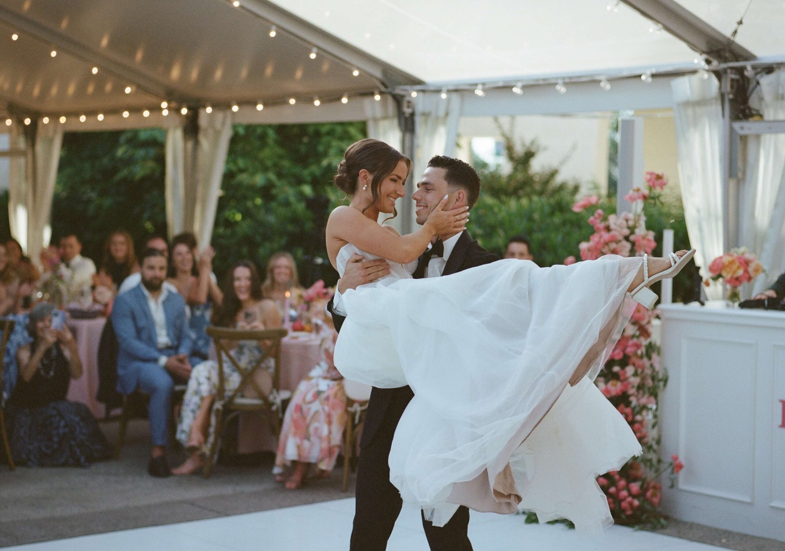 A bride and groom share their first dance under a tent adorned with string lights. Guests are seated at tables decorated with flowers, watching them.
