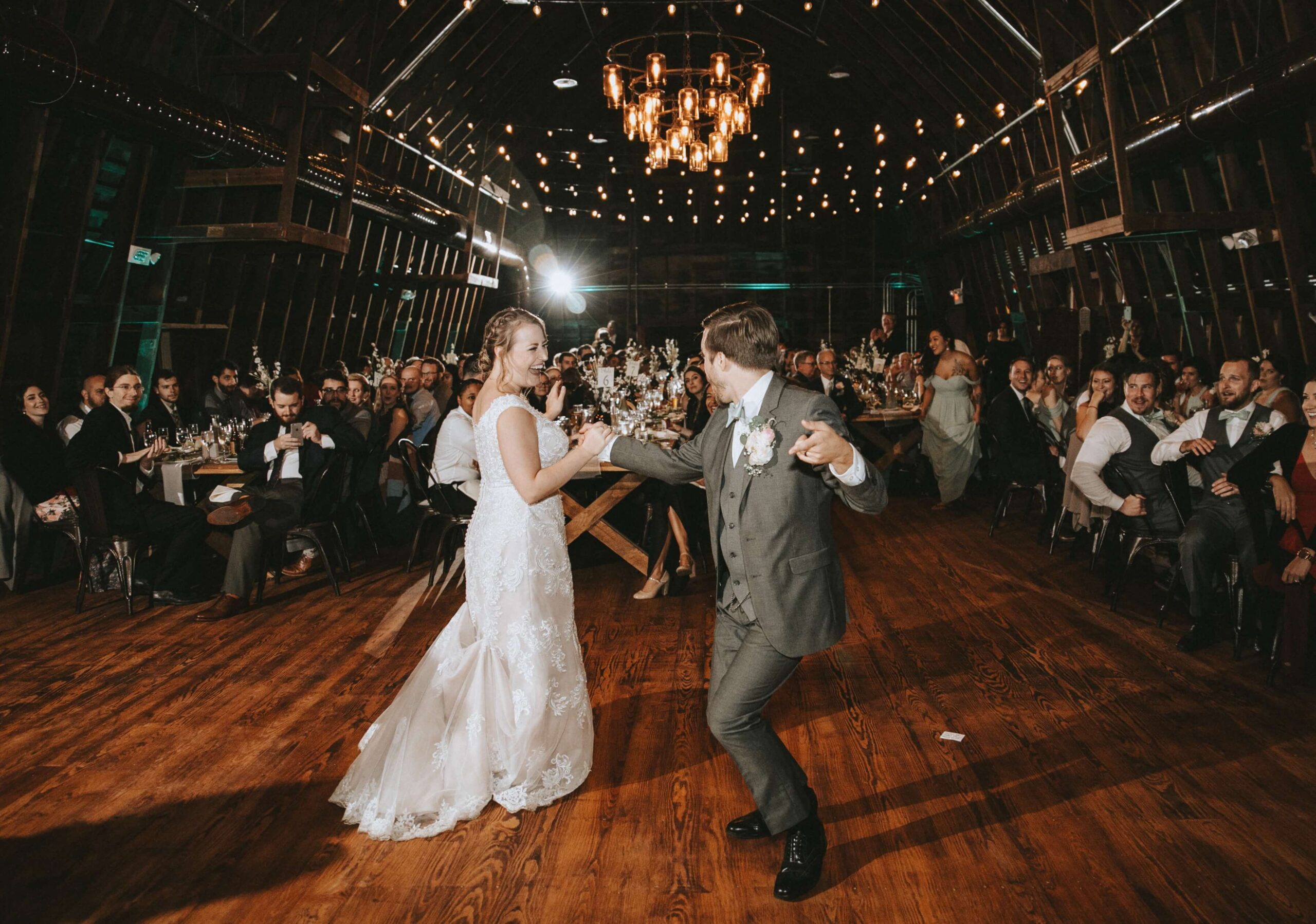 A couple is dancing at a wedding reception in a rustic barn setting, surrounded by seated guests.