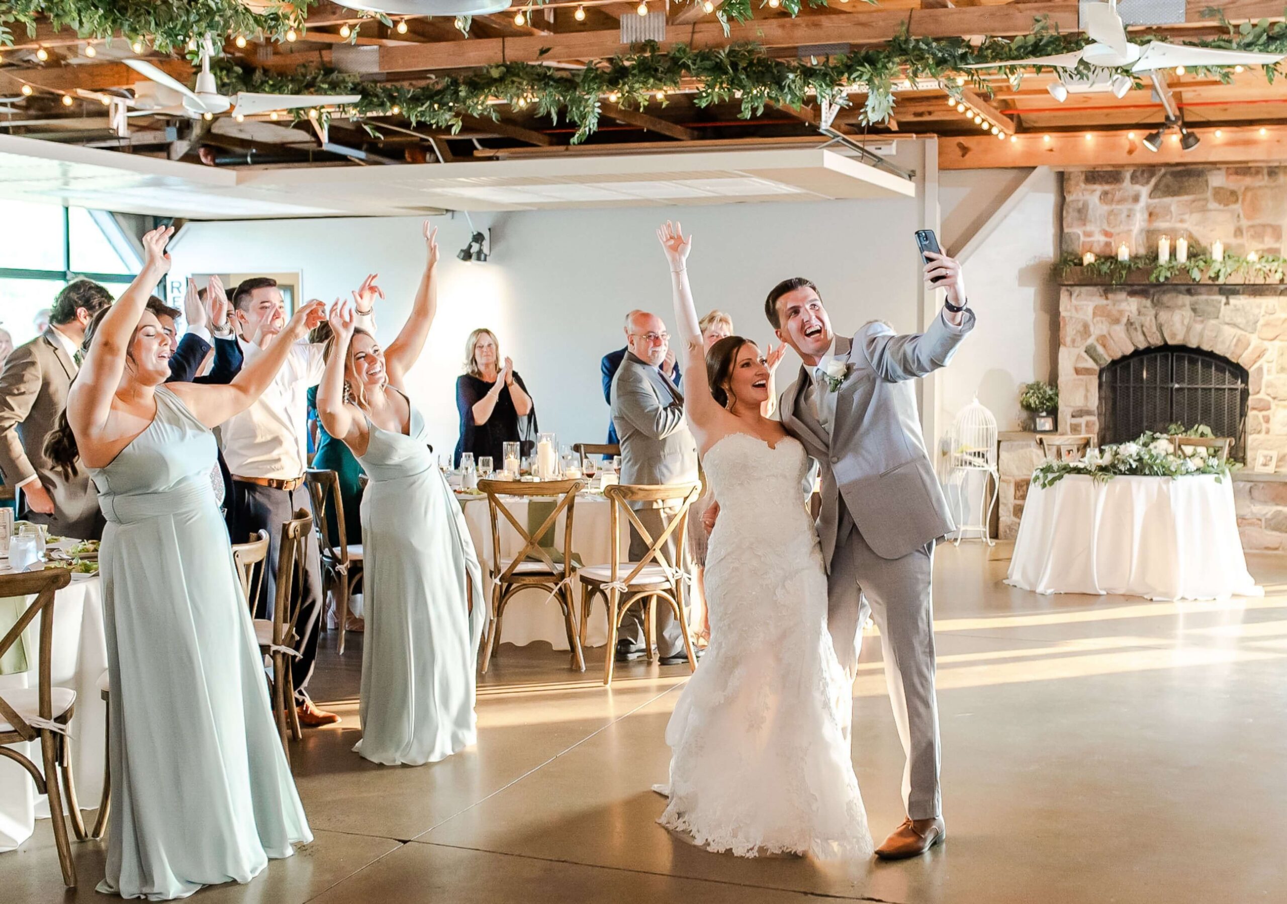 A bride and groom take a selfie while guests cheer. The room is decorated with greenery and has a stone fireplace.