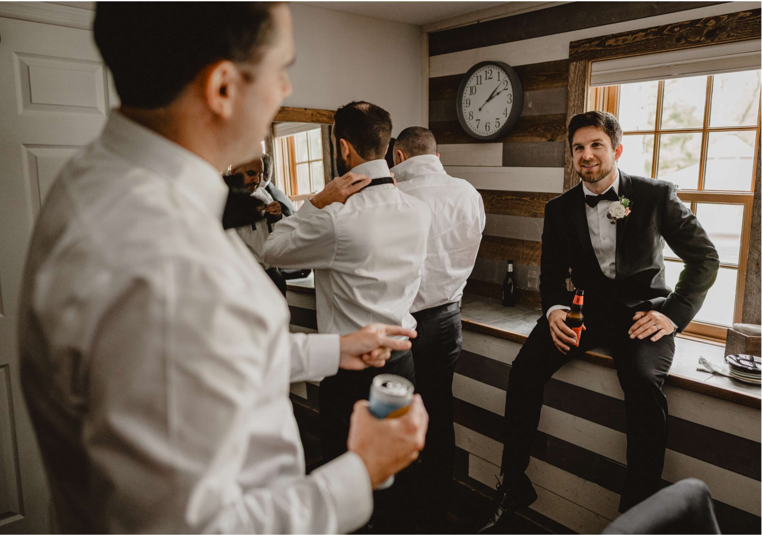 Groom and groomsmen in a room getting ready. One man adjusts his tie by a mirror, another holds a drink, and the groom, holding a beer, sits on a ledge. A clock is on the wall.