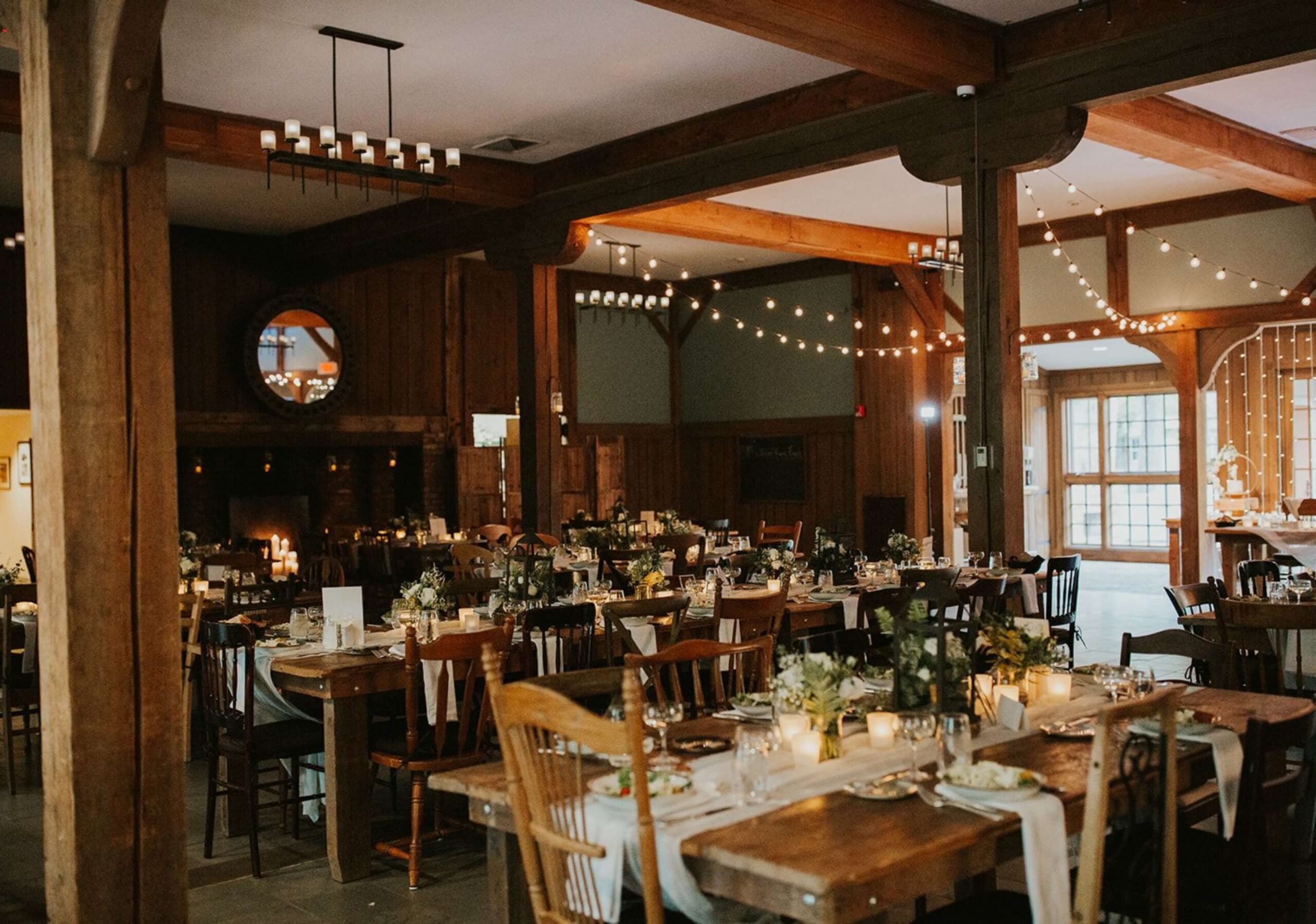 A rustic banquet hall with wooden beams is set for an event. Tables are decorated with greenery, candles, and tableware. String lights hang from the ceiling, creating a warm ambiance.