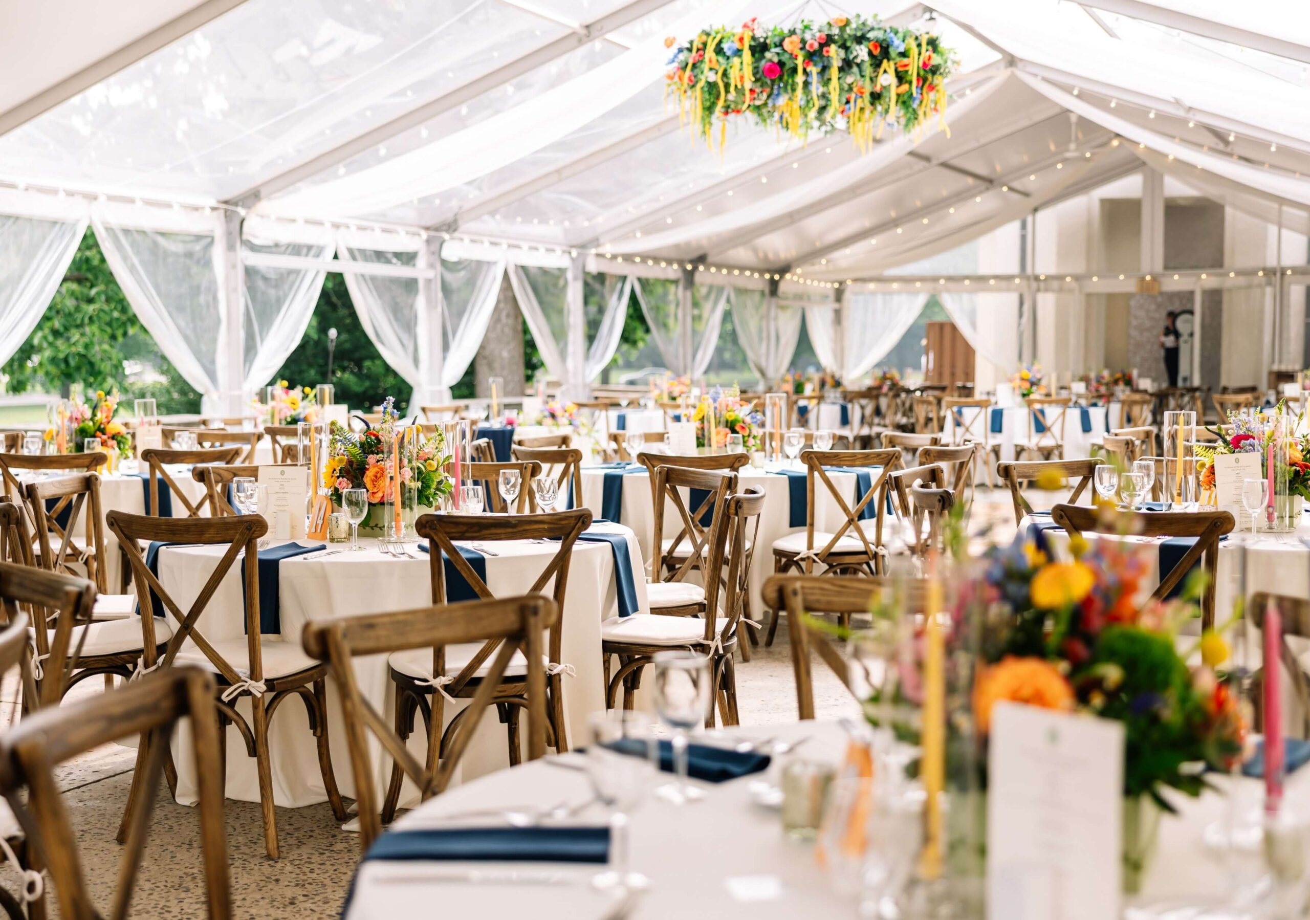 Elegant outdoor event setup under a clear tent with wooden chairs and round tables adorned with colorful floral centerpieces and blue accents.