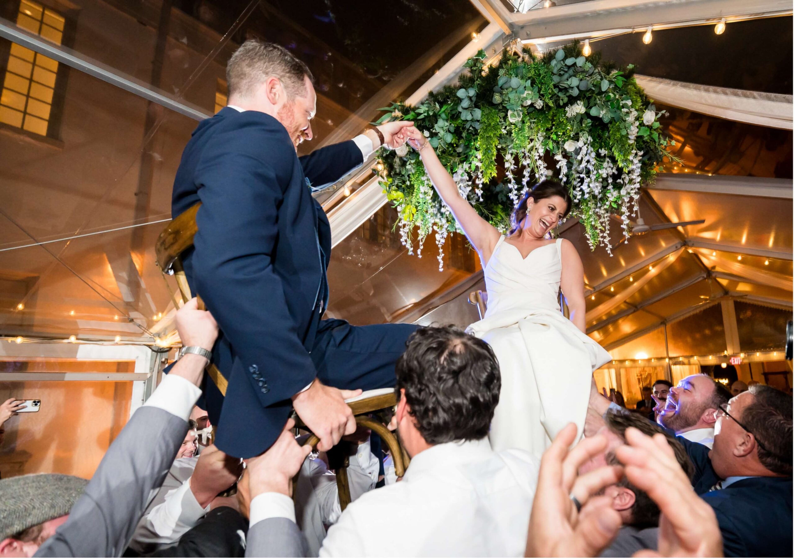 A bride and groom are lifted on chairs by guests during a wedding celebration under a tent with hanging greenery.