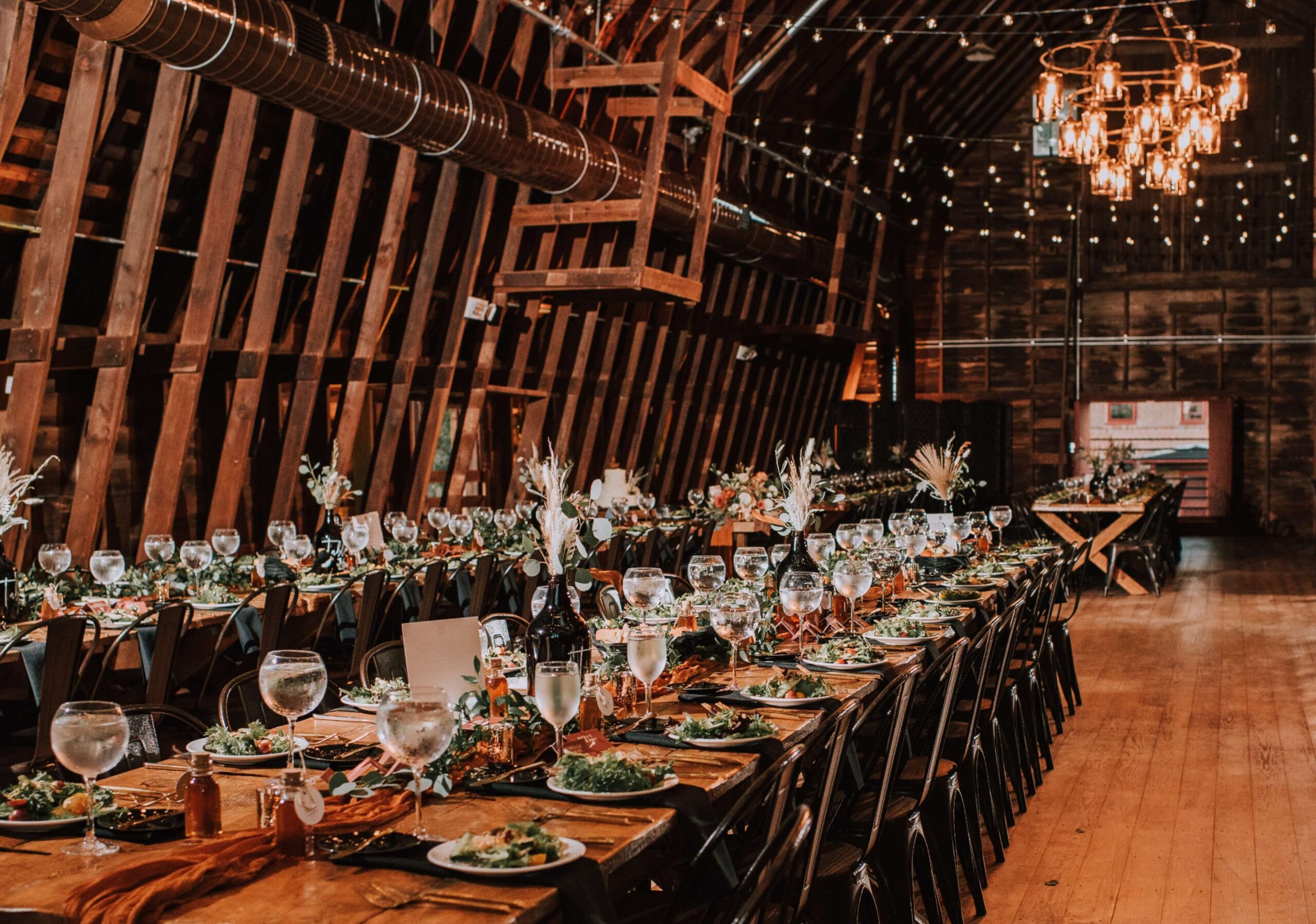 Rustic barn interior with long dining tables set for an event. Tables feature salads, wine glasses, and small decor. Chandeliers and string lights hang from the wooden ceiling.