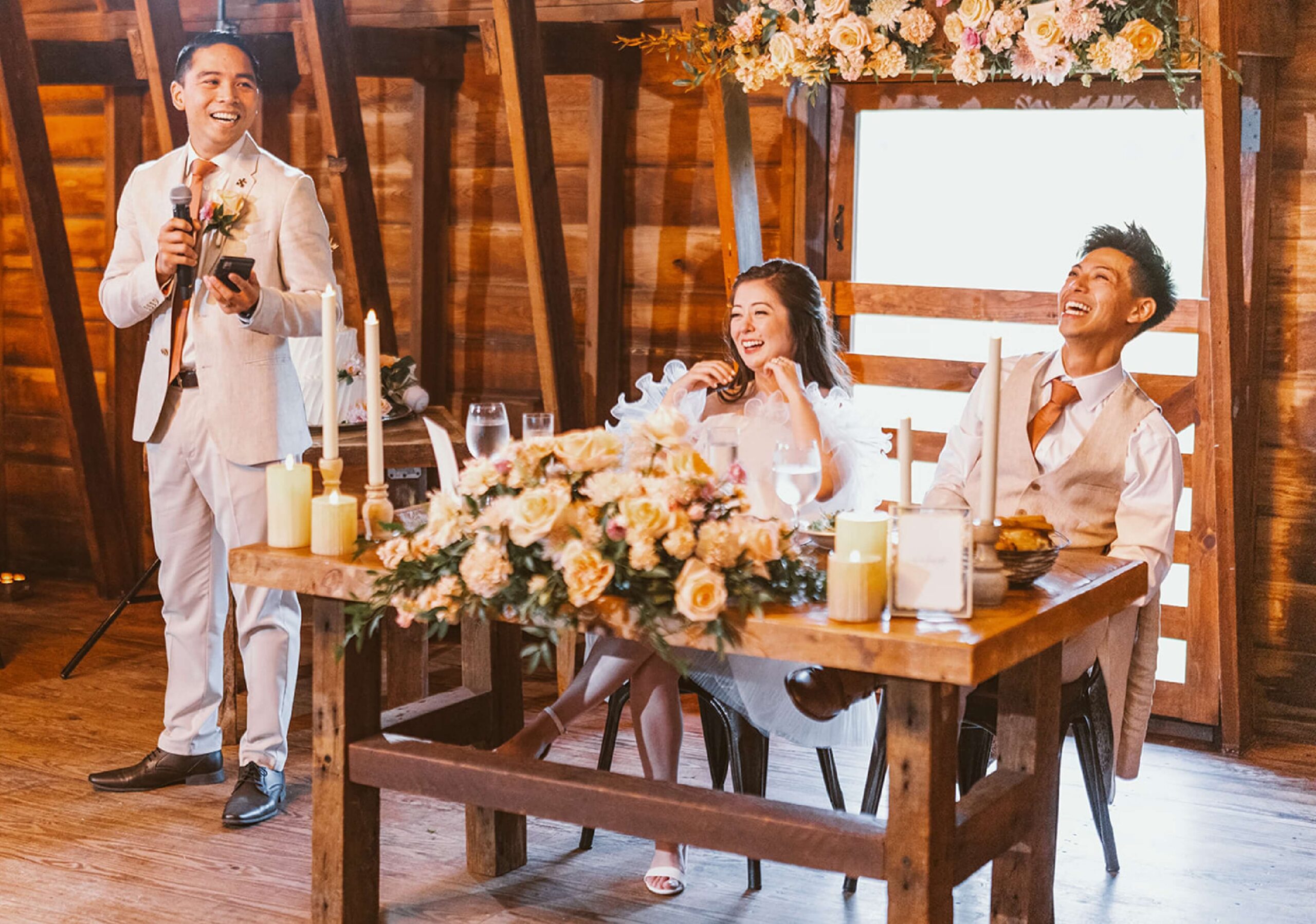 A man speaks into a microphone standing next to a seated couple at a wooden table adorned with candles and flowers, all dressed in formal attire, in a rustic wooden venue.