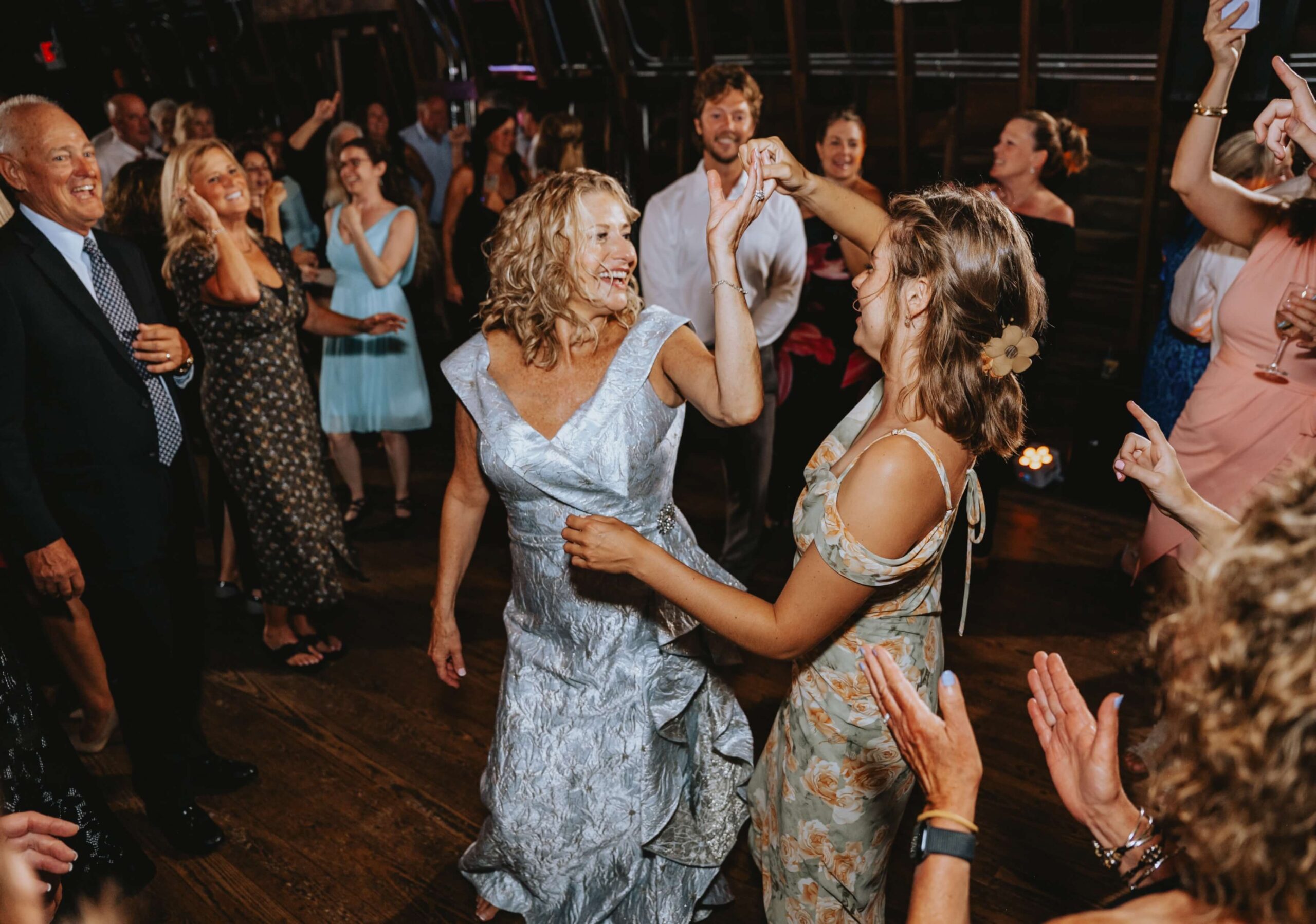 Two women dance joyfully together at a lively indoor party, surrounded by cheering people.