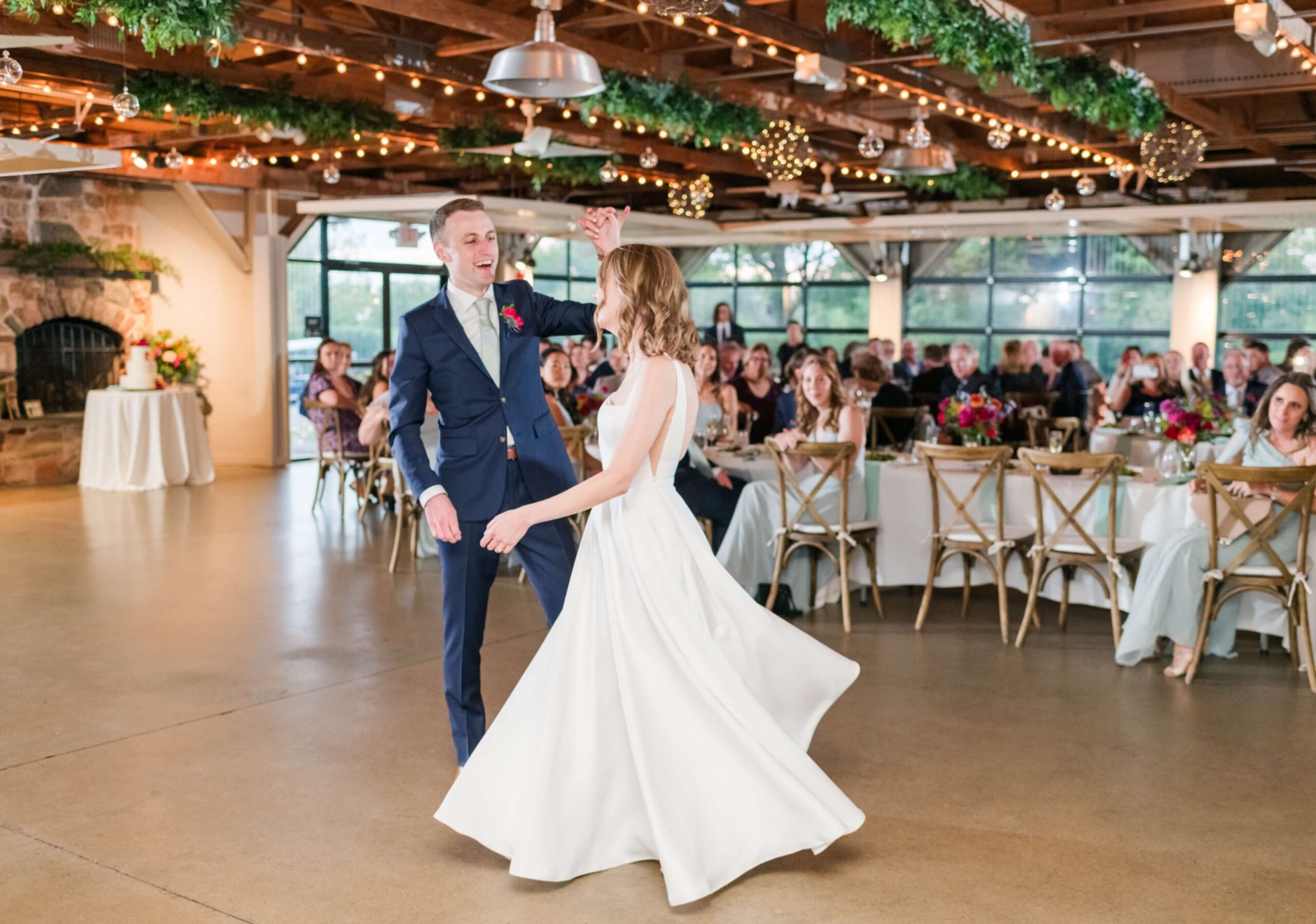 A couple in formal attire dances in a decorated reception hall with guests seated at round tables in the background.