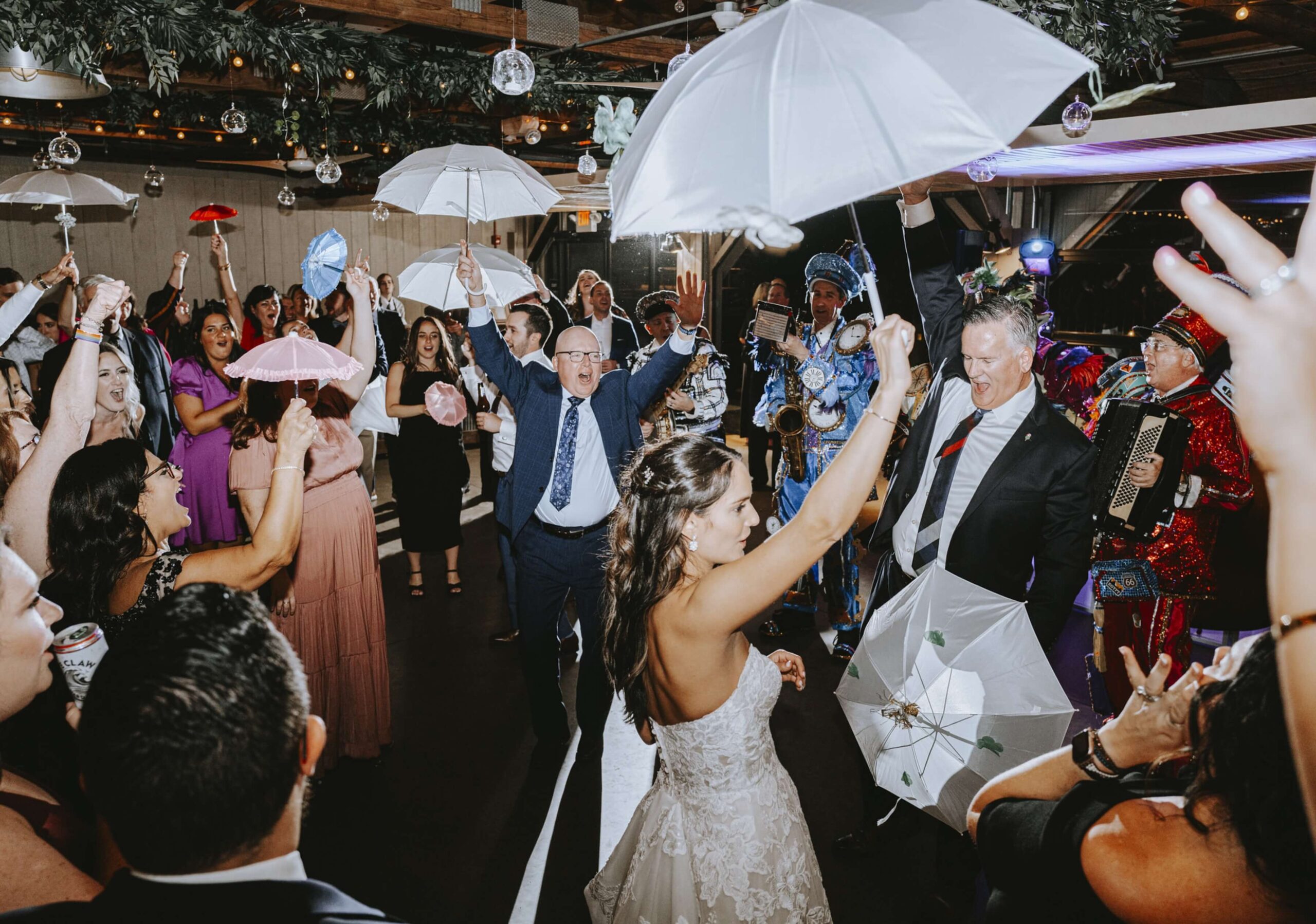 A bride and guests are dancing at a wedding reception, holding white umbrellas. The group is celebrating under hanging decorations in a lively atmosphere.