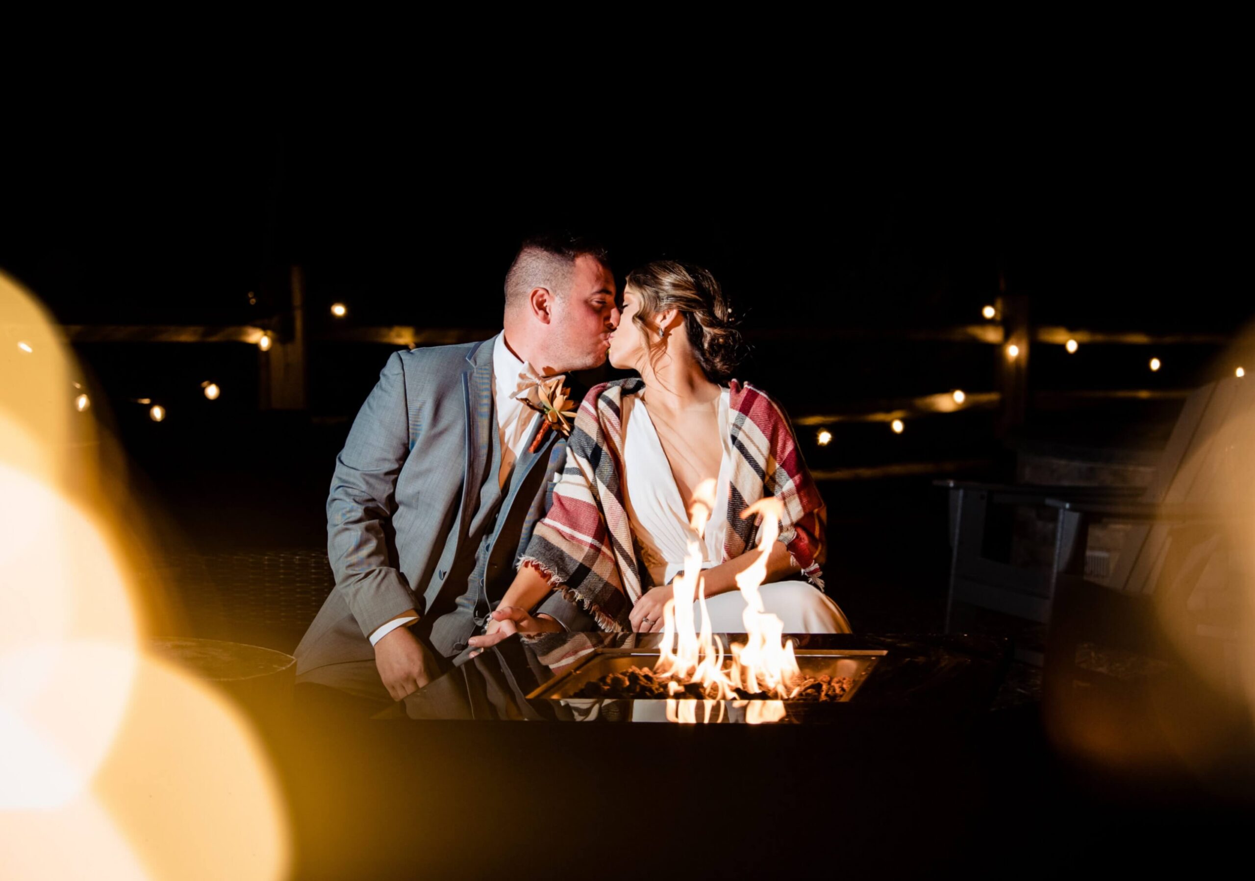A couple in formal attire sits by a fire pit, kissing under string lights at night. The woman is wrapped in a plaid shawl.