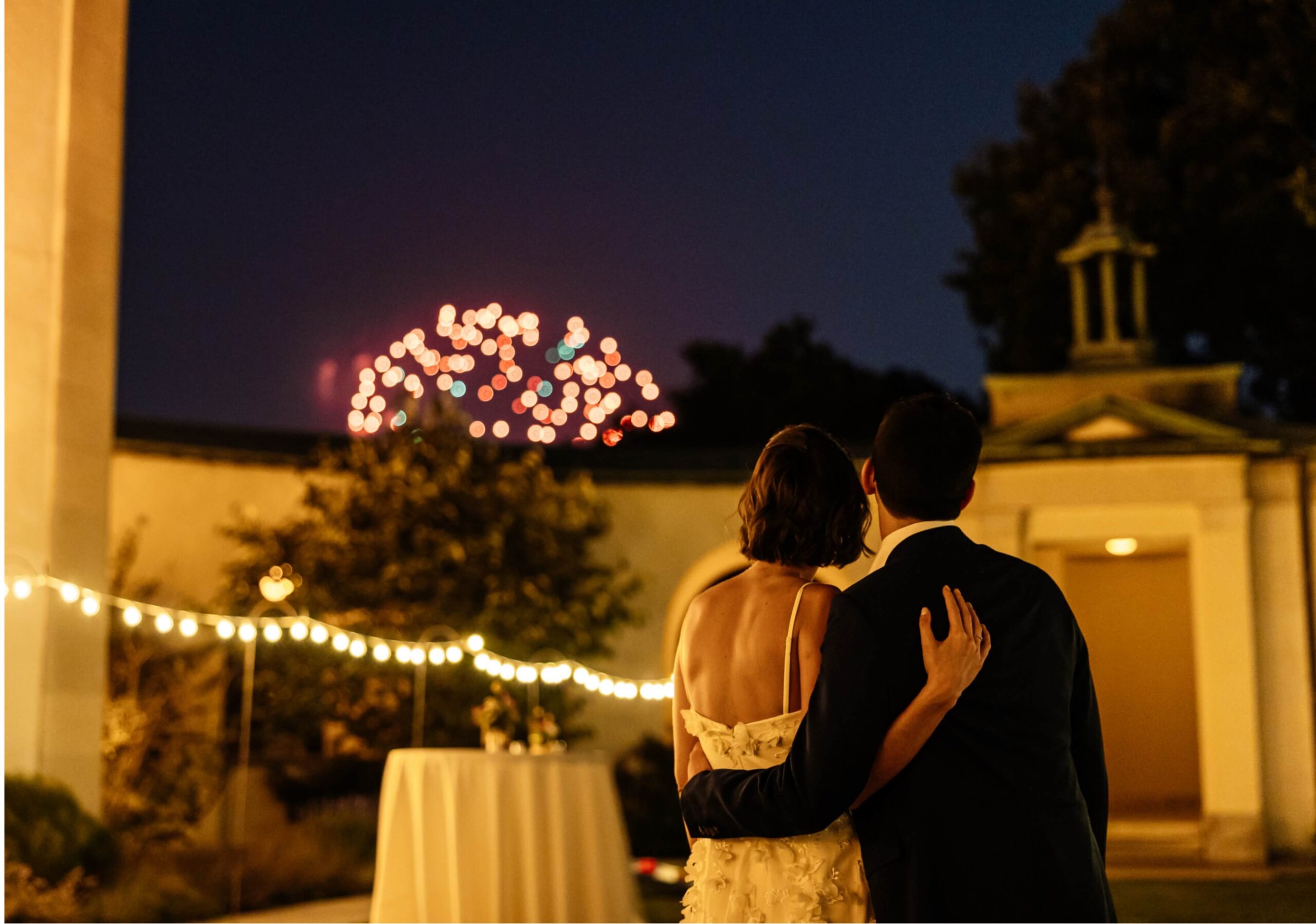 A couple embraces while watching fireworks in the night sky, with string lights and a building in the background.