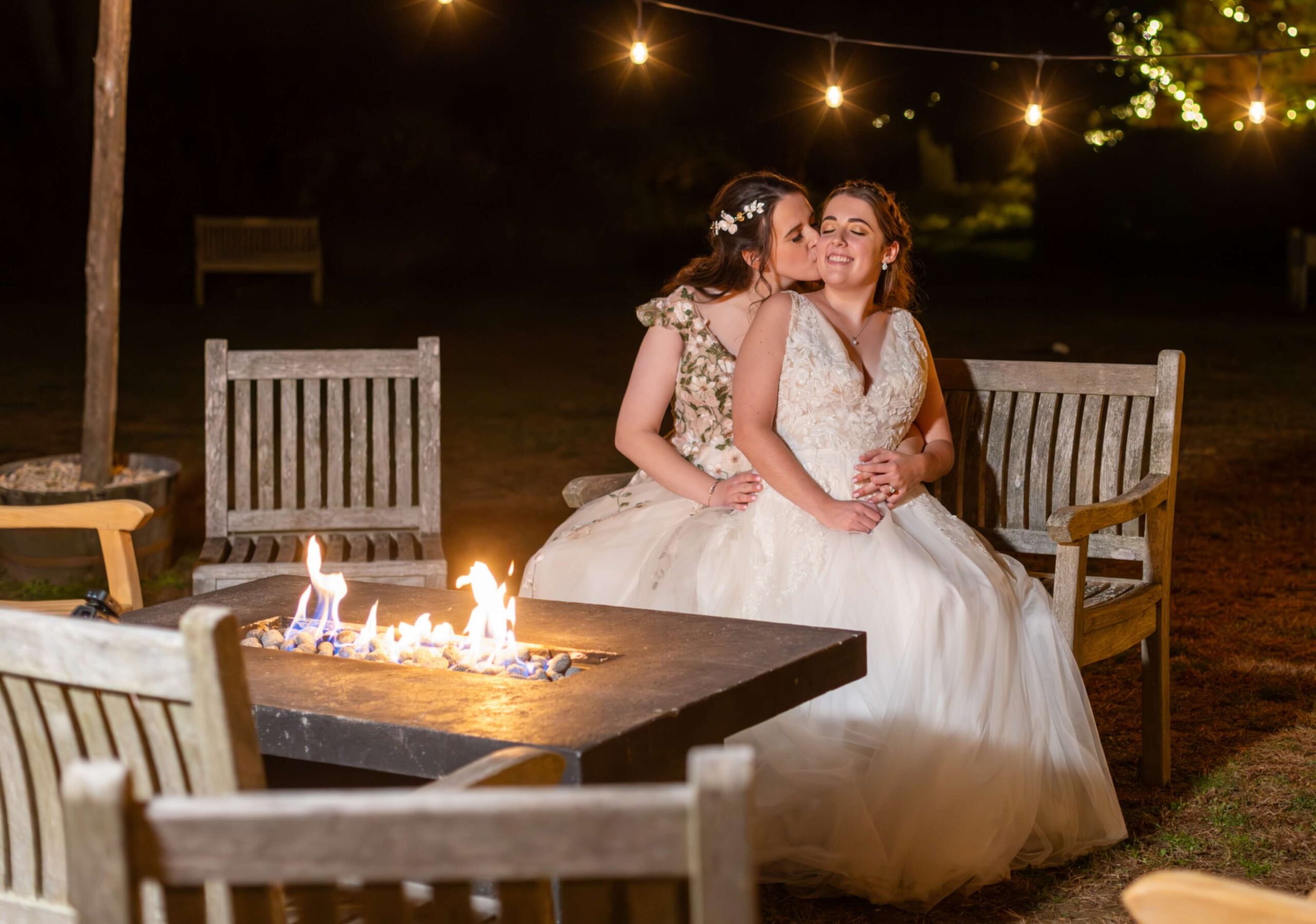 Two women in wedding dresses share an embrace on a wooden bench by a lit fire pit, with string lights overhead at night.