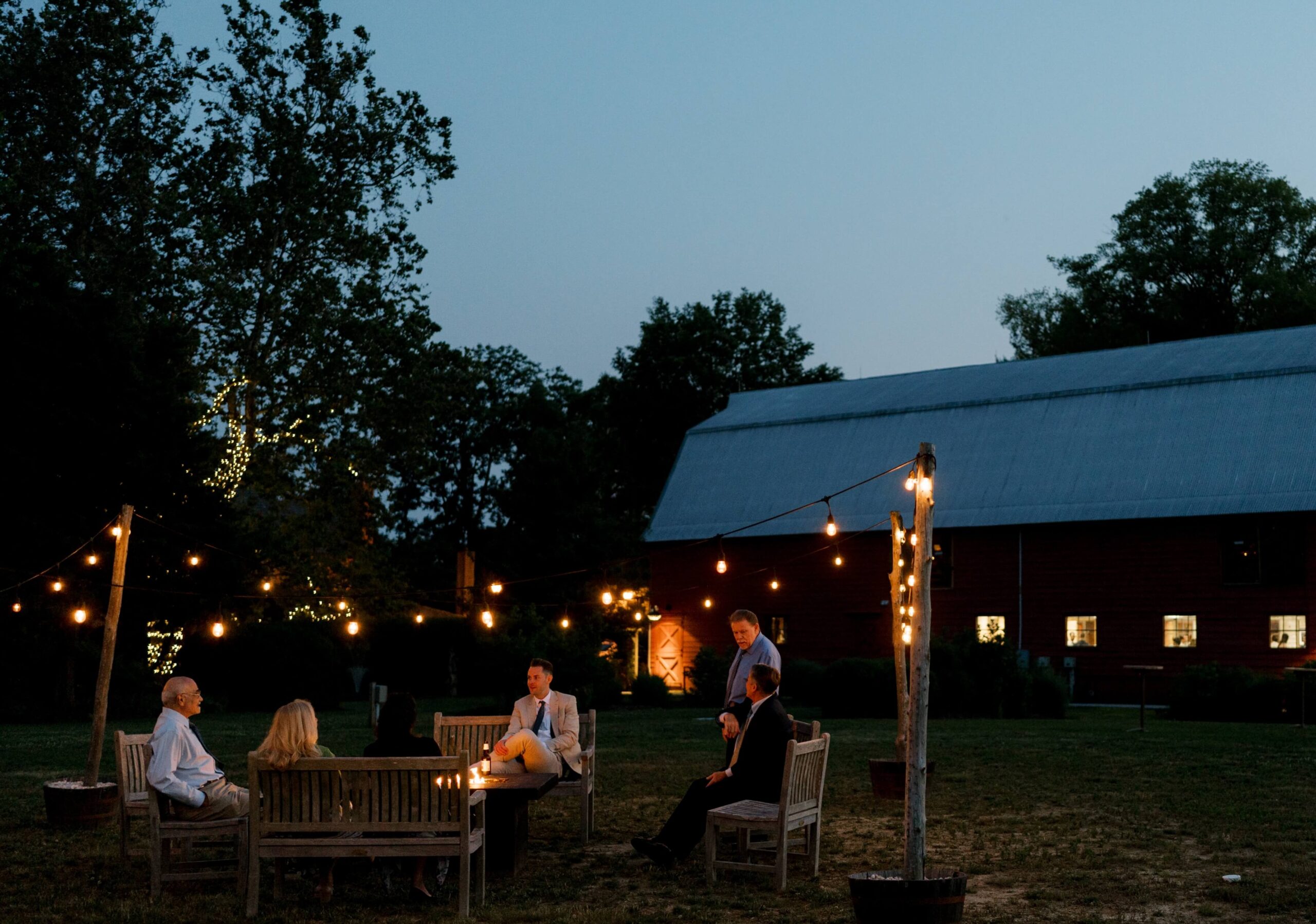 People sit around a fire pit on wooden chairs in a grassy area at dusk. A barn with windows is lit up in the background, with string lights hanging overhead.