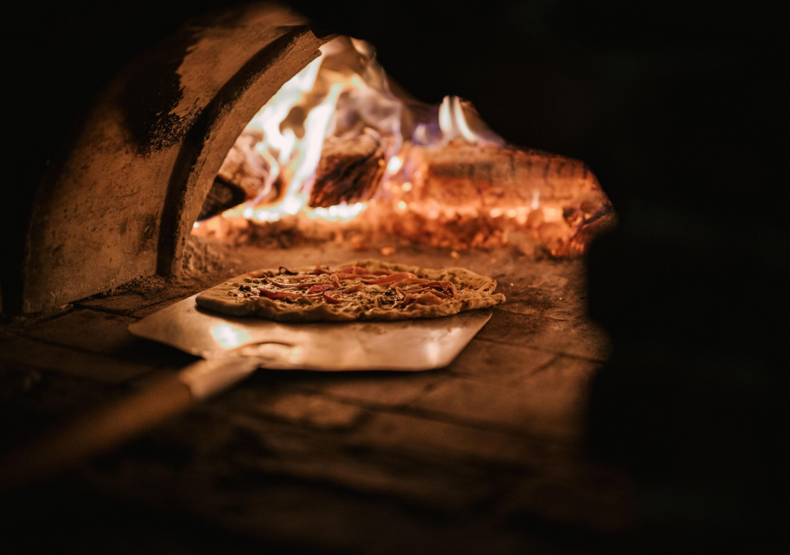 A pizza being placed in a wood-fired oven with visible flames and glowing embers.