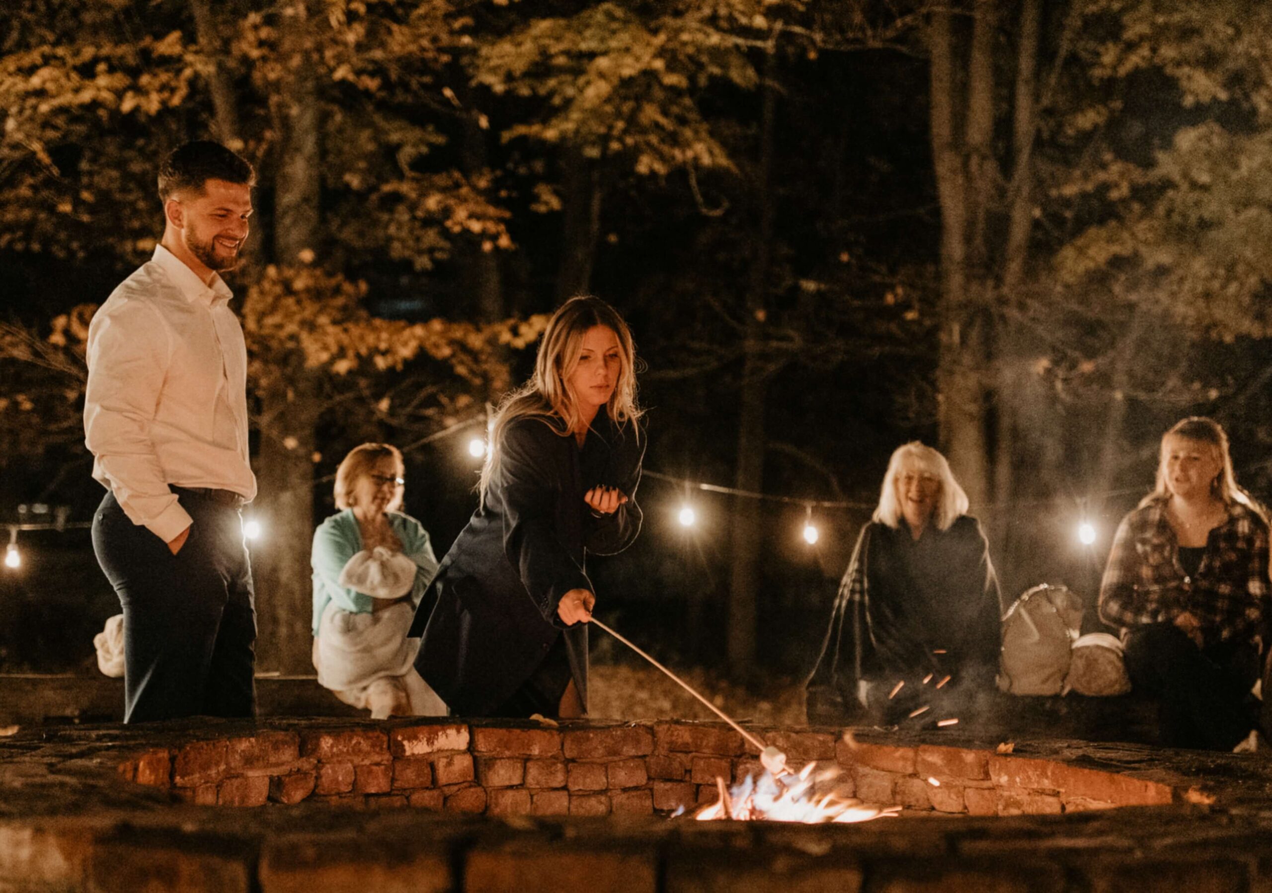 People gathered around a brick fire pit at night, with one person roasting a marshmallow. String lights and trees are in the background.