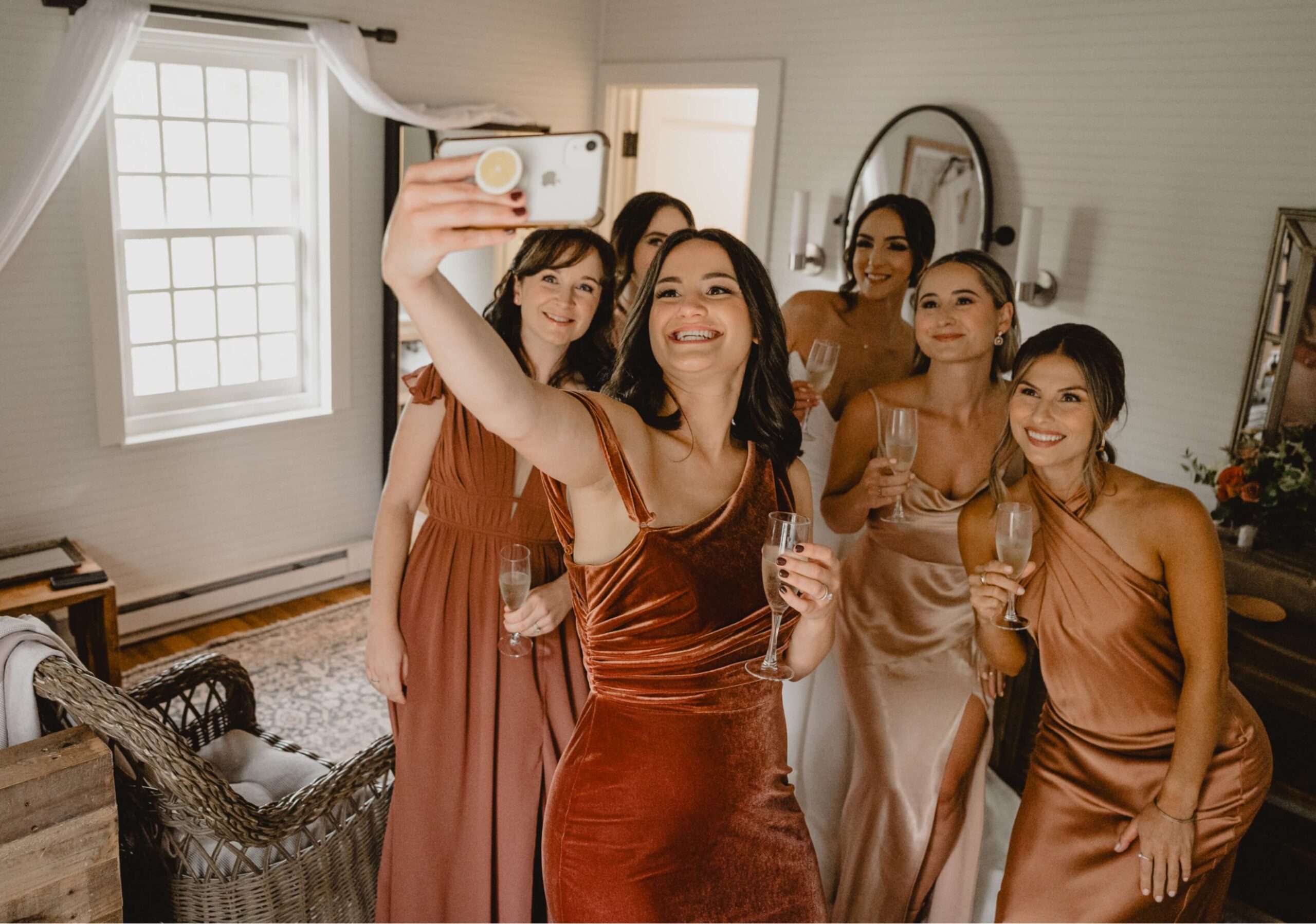 A group of six women in formal dresses take a selfie in a well-lit room, holding champagne glasses.