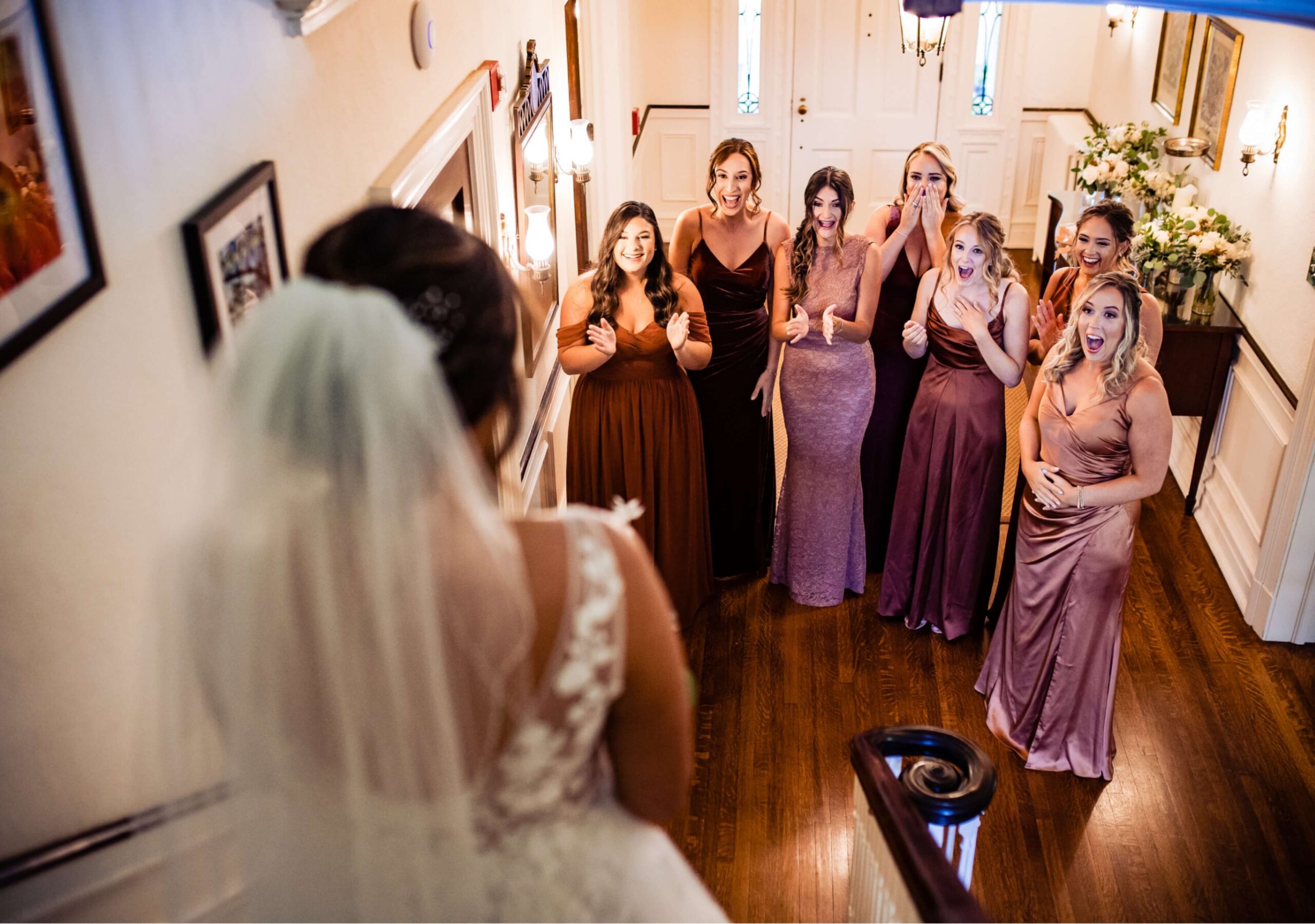 A bride stands at the top of a staircase while a group of seven bridesmaids in different brown dresses react with excitement and surprise.