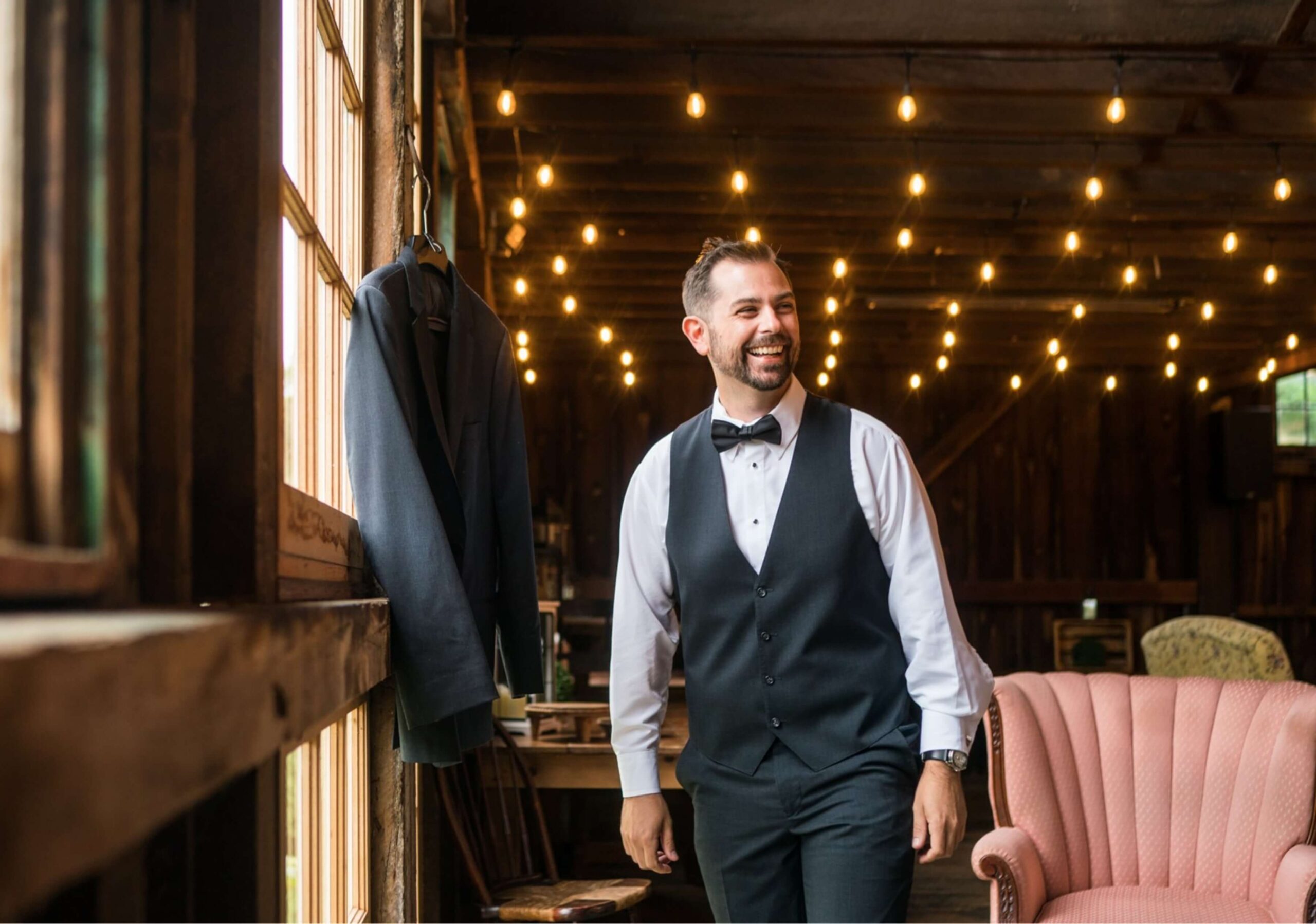 Man in a tuxedo vest smiles, standing indoors with string lights above. A suit jacket hangs on a window next to a pink sofa.