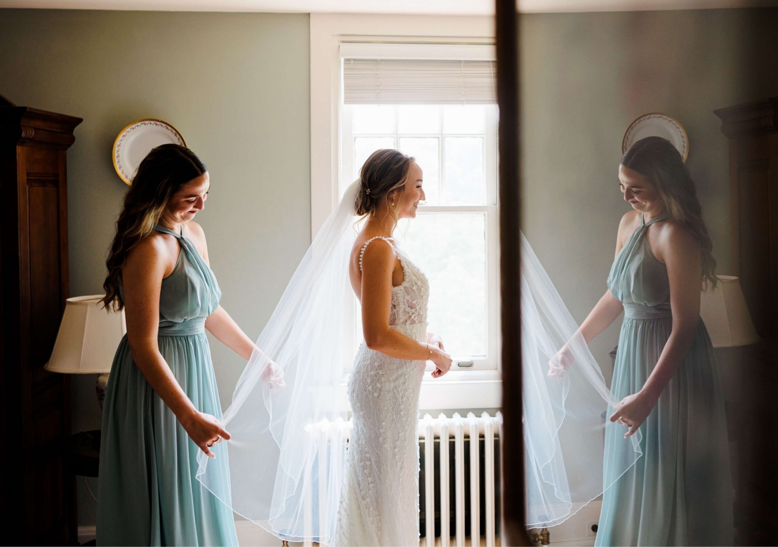 Bride in white dress and veil stands by a window, smiling, while a bridesmaid in a light blue dress adjusts the veil in a softly lit room.