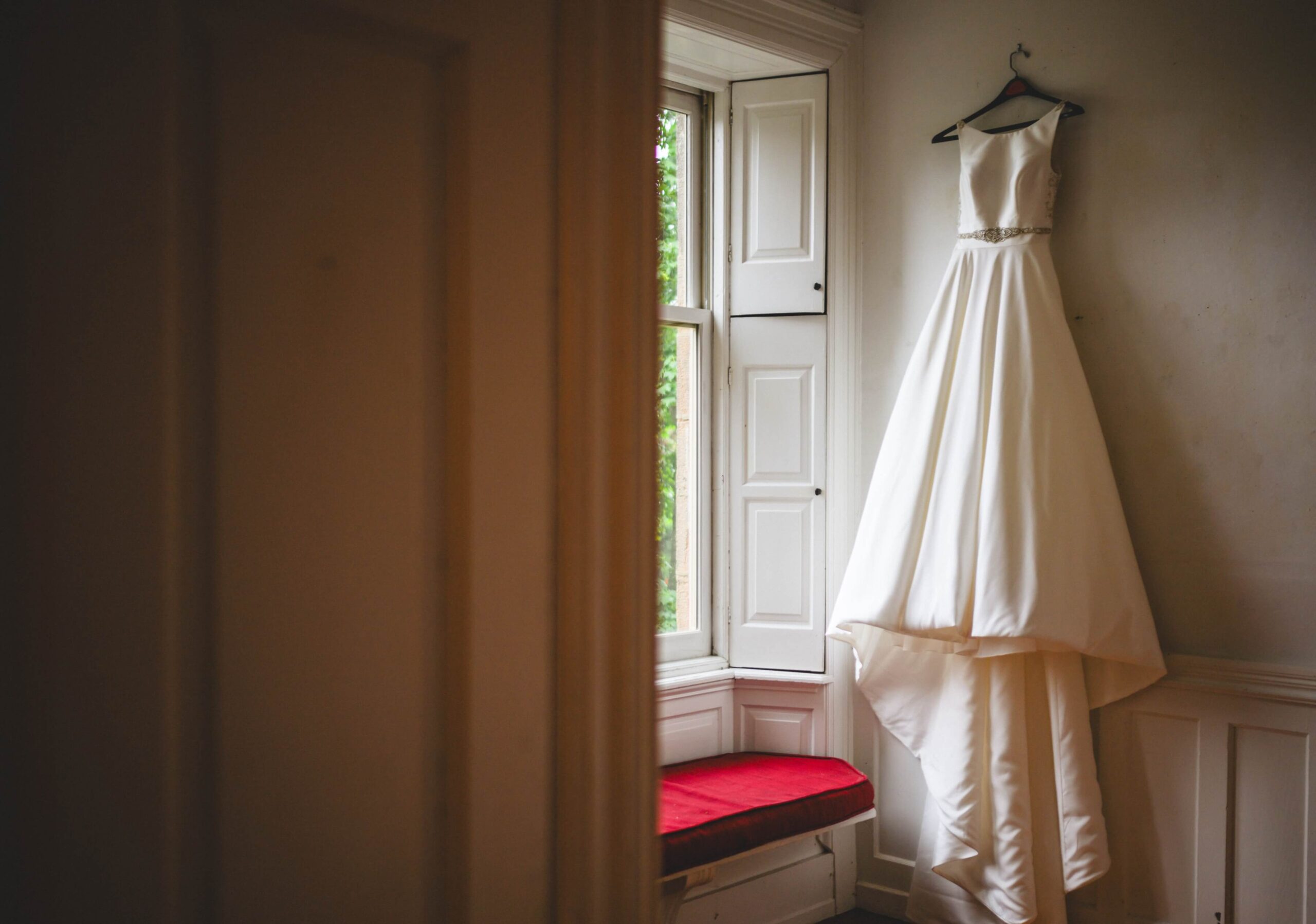 A white wedding dress hangs on a hanger in front of a window with red-cushioned seating.
