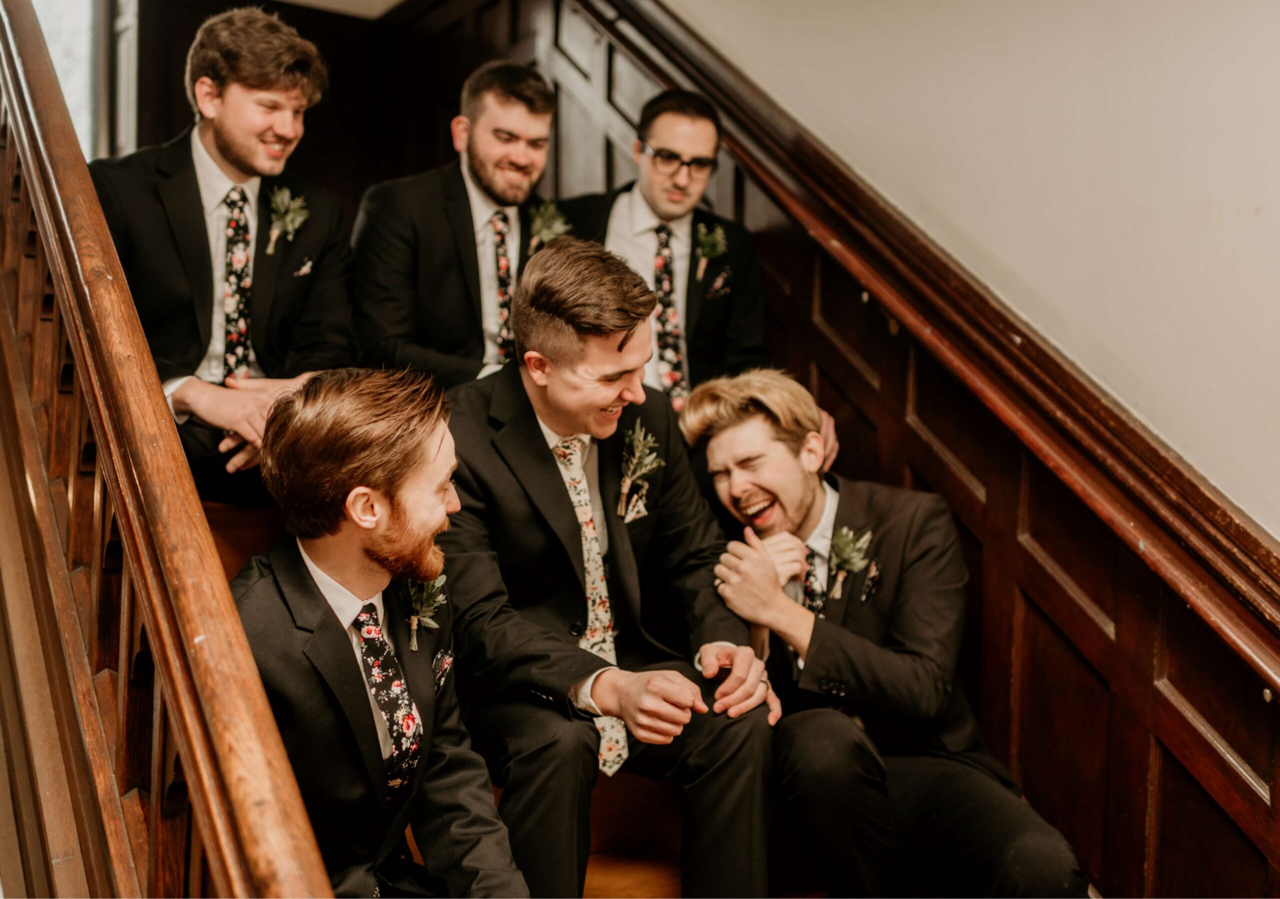 Six men in suits sit and laugh on a wooden staircase.