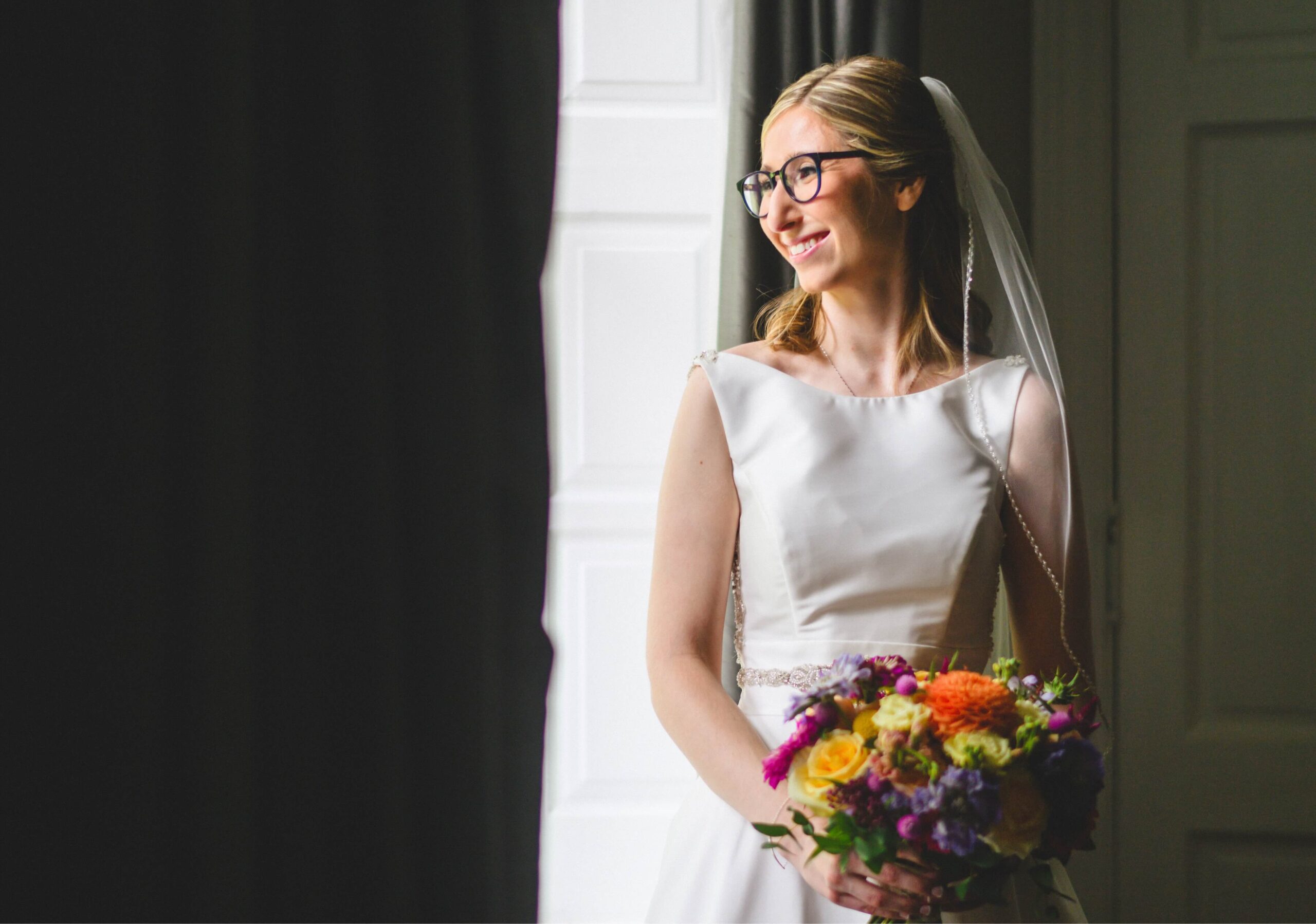 Bride in a white dress and veil stands smiling, holding a vibrant bouquet of flowers, near a large window with white curtains.