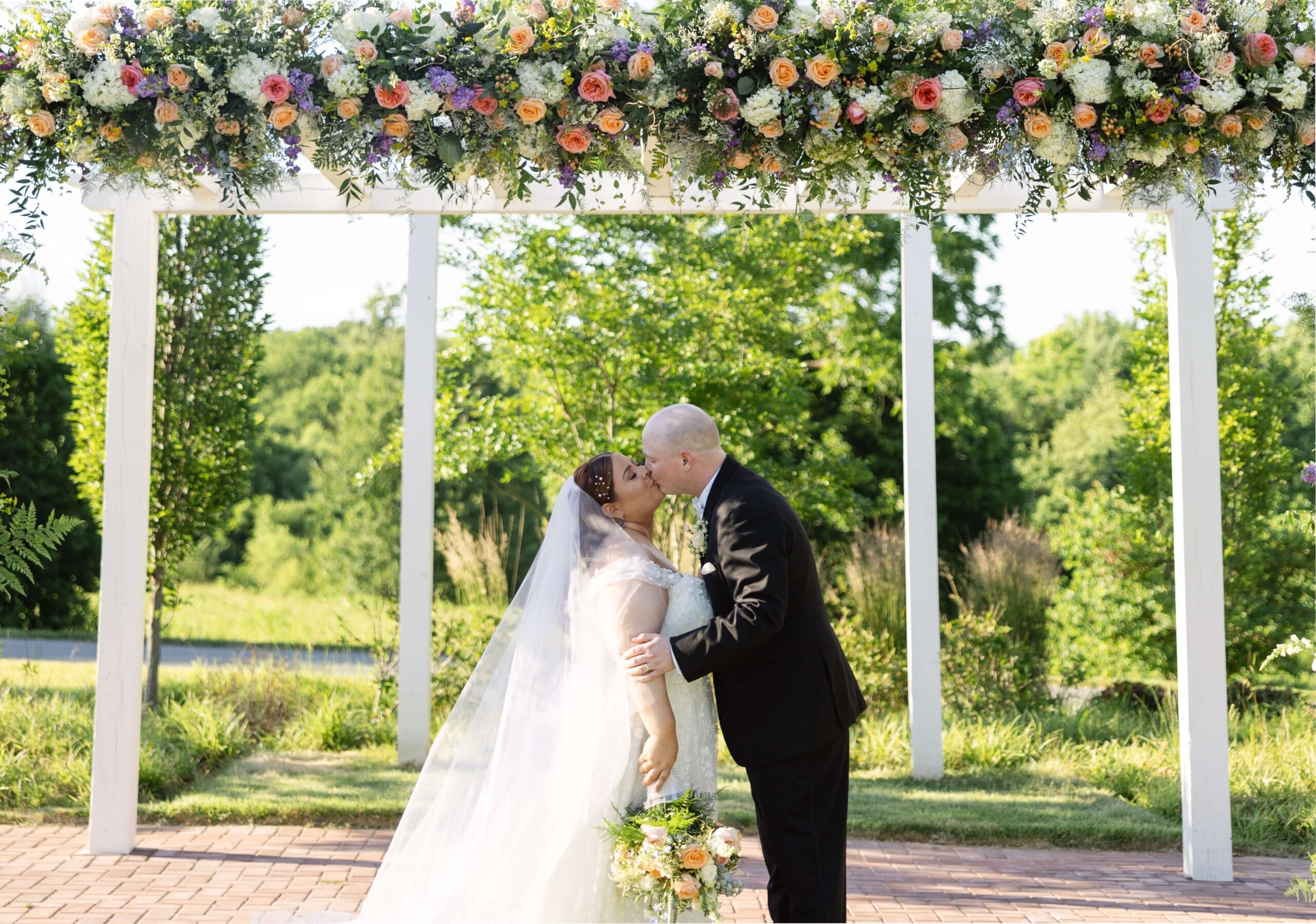 Bride and groom kissing under a flower-covered pergola in an outdoor setting with greenery in the background.