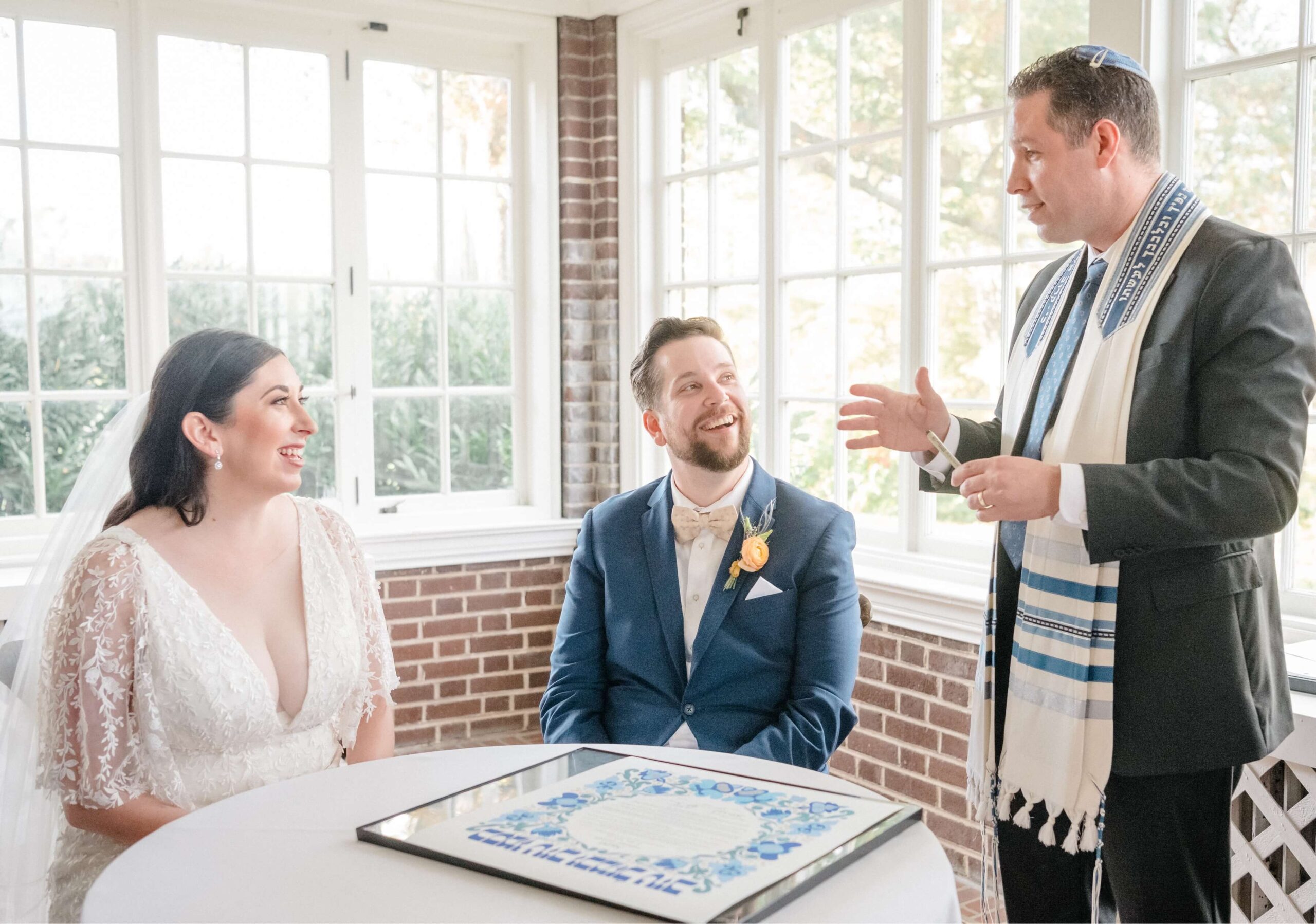 A couple in wedding attire sits at a table with a document, while a man standing beside them gestures as he speaks. The room is bright with large windows in the background.