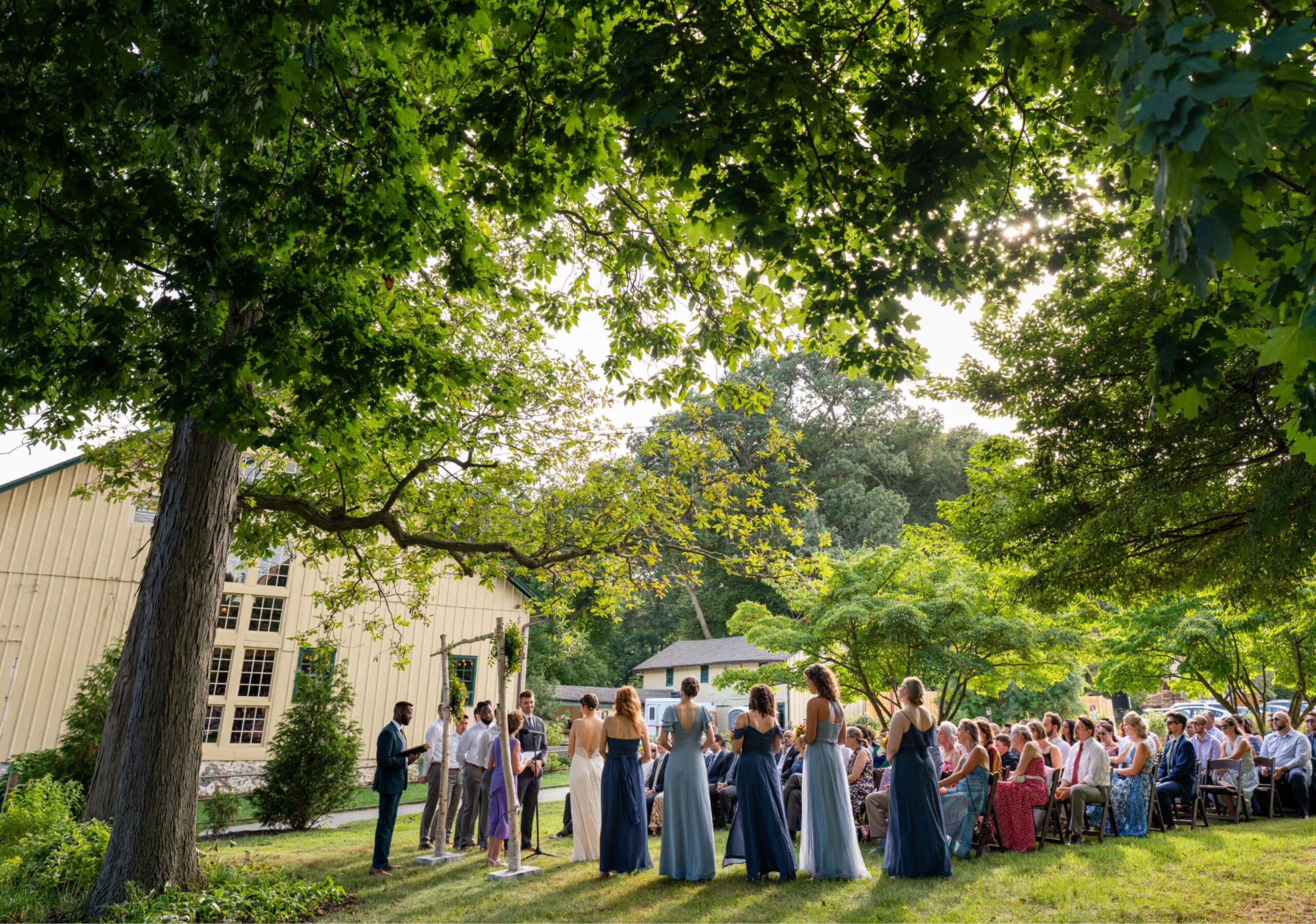 Outdoor wedding ceremony with guests seated on chairs under trees near a large building, with a couple standing at the altar surrounded by the wedding party.