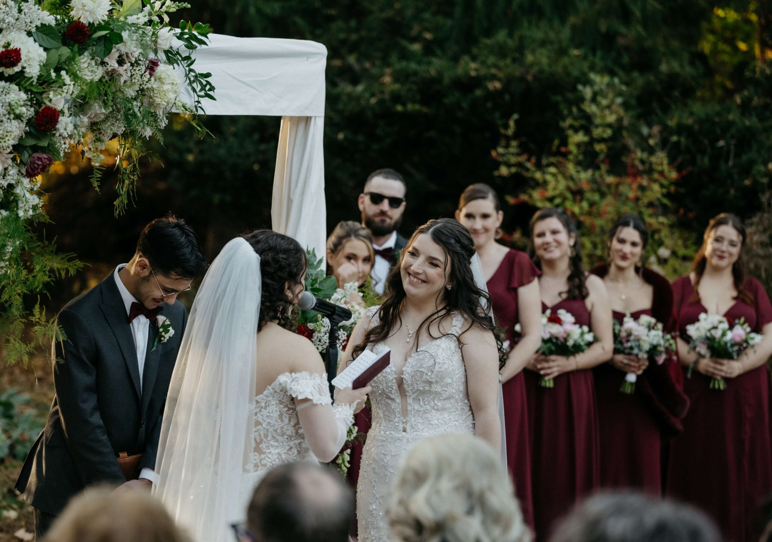 Two brides stand under an arch during an outdoor wedding ceremony. One reads vows from paper. Bridesmaids in burgundy dresses stand in the background.