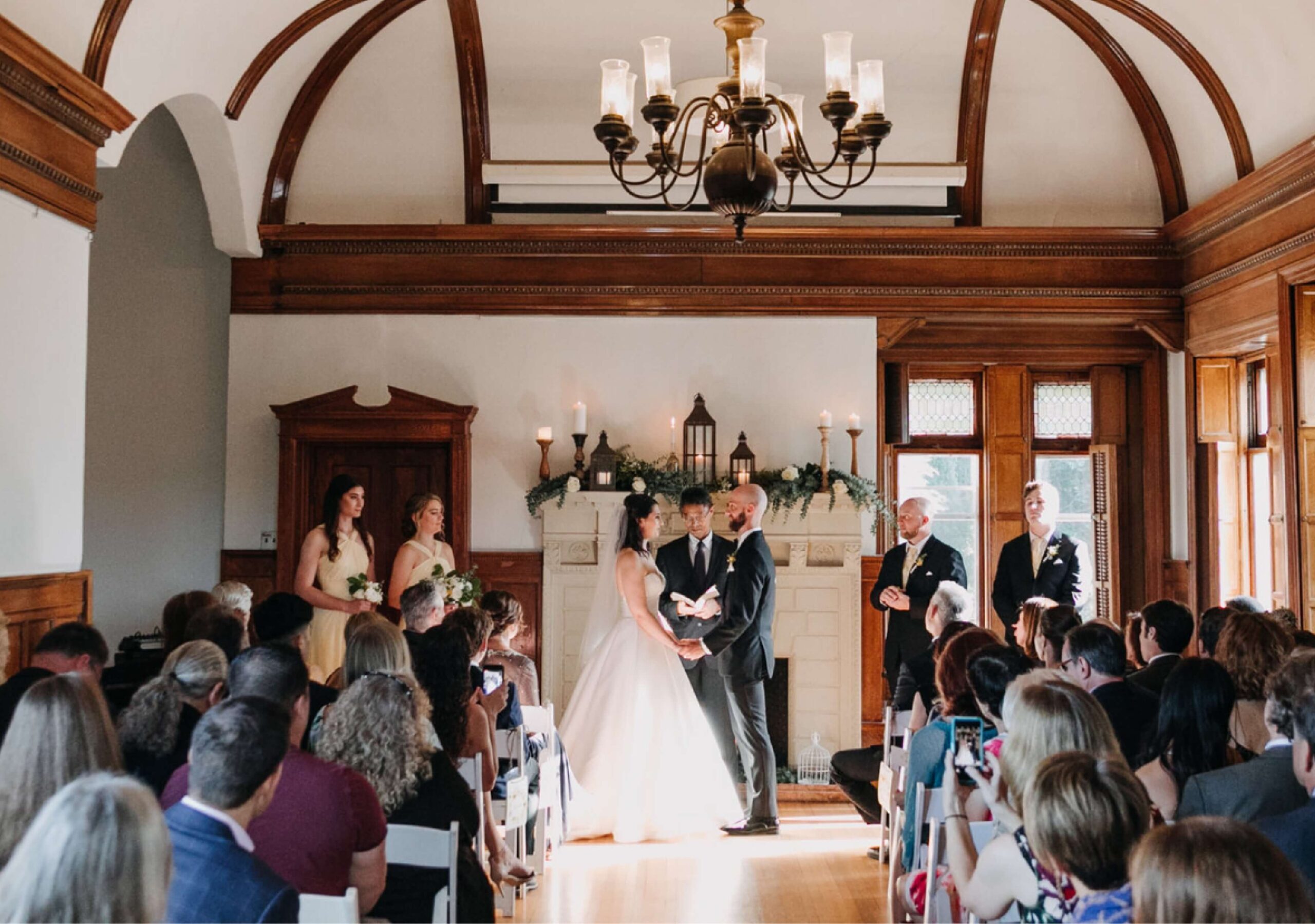A bride and groom stand at an indoor wedding ceremony with a small audience seated on both sides. Bridesmaids and groomsmen are positioned nearby. The room features wood accents and arched ceilings.