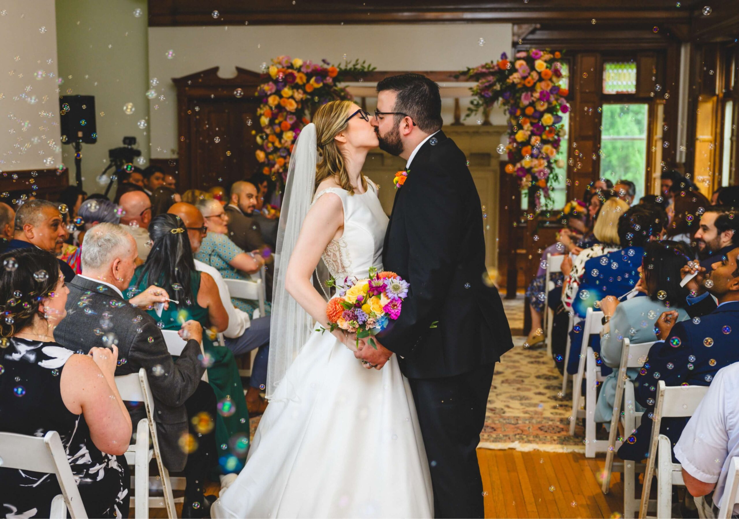 A bride and groom kiss at the end of a wedding aisle. They hold a bouquet, surrounded by guests seated in rows. Bubbles float in the air, and a floral arch is visible in the background.
