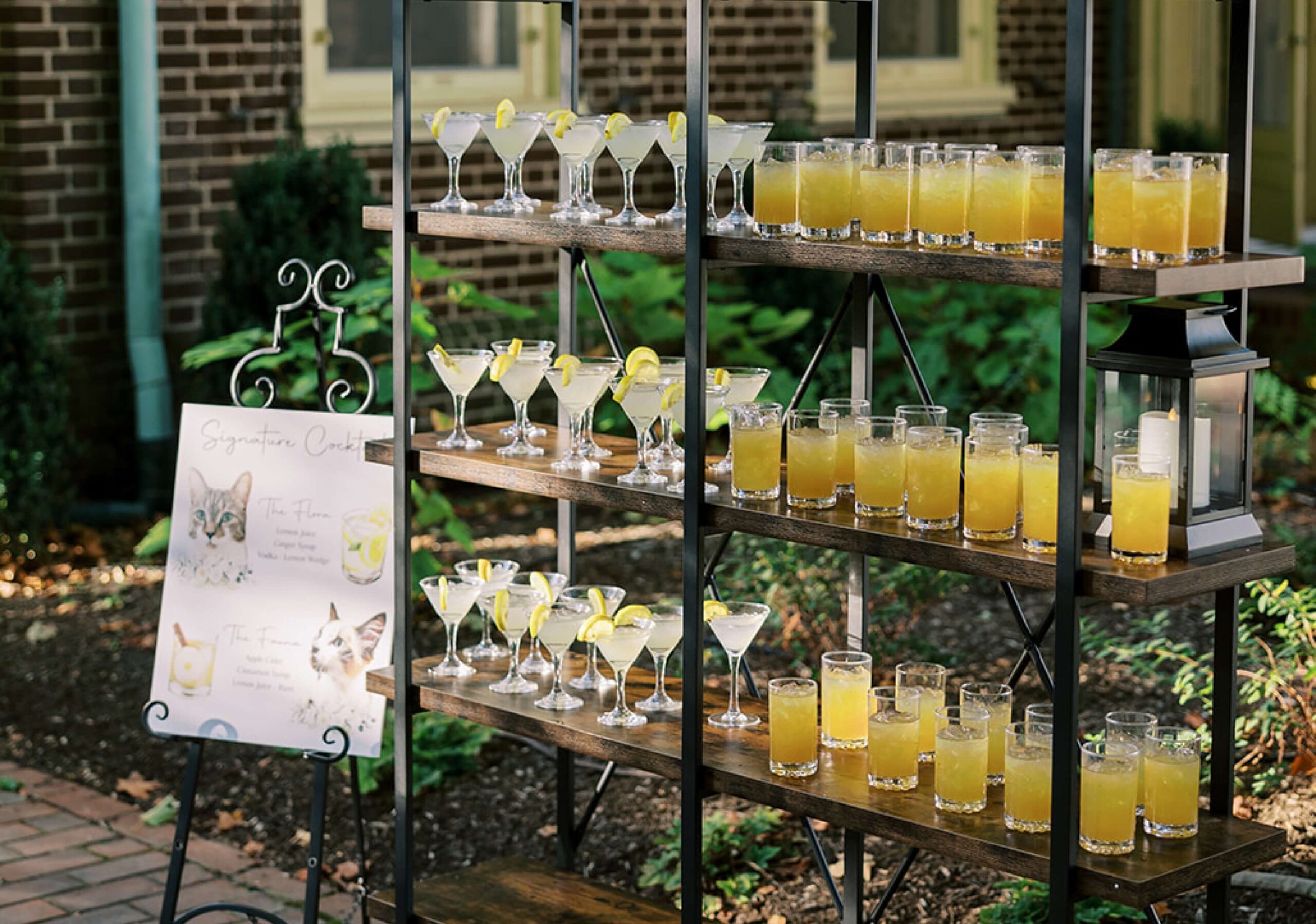 A display of various cocktails on three wooden shelves, with a standing menu sign beside them. Some drinks have lemon garnishes. Brick wall and foliage in the background.