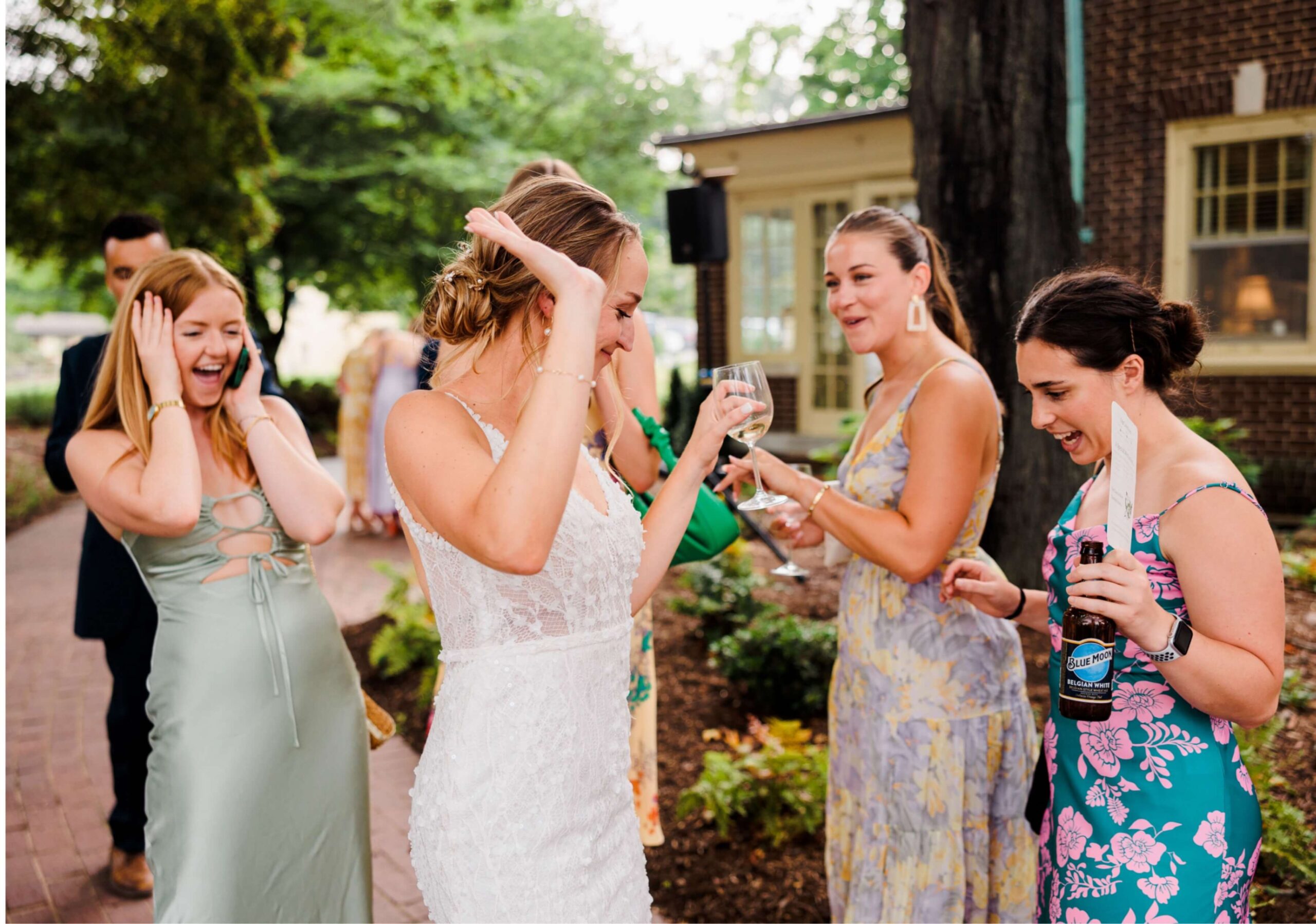A bride in a white dress laughs with three women holding drinks, outdoors on a pathway with trees and a building in the background.