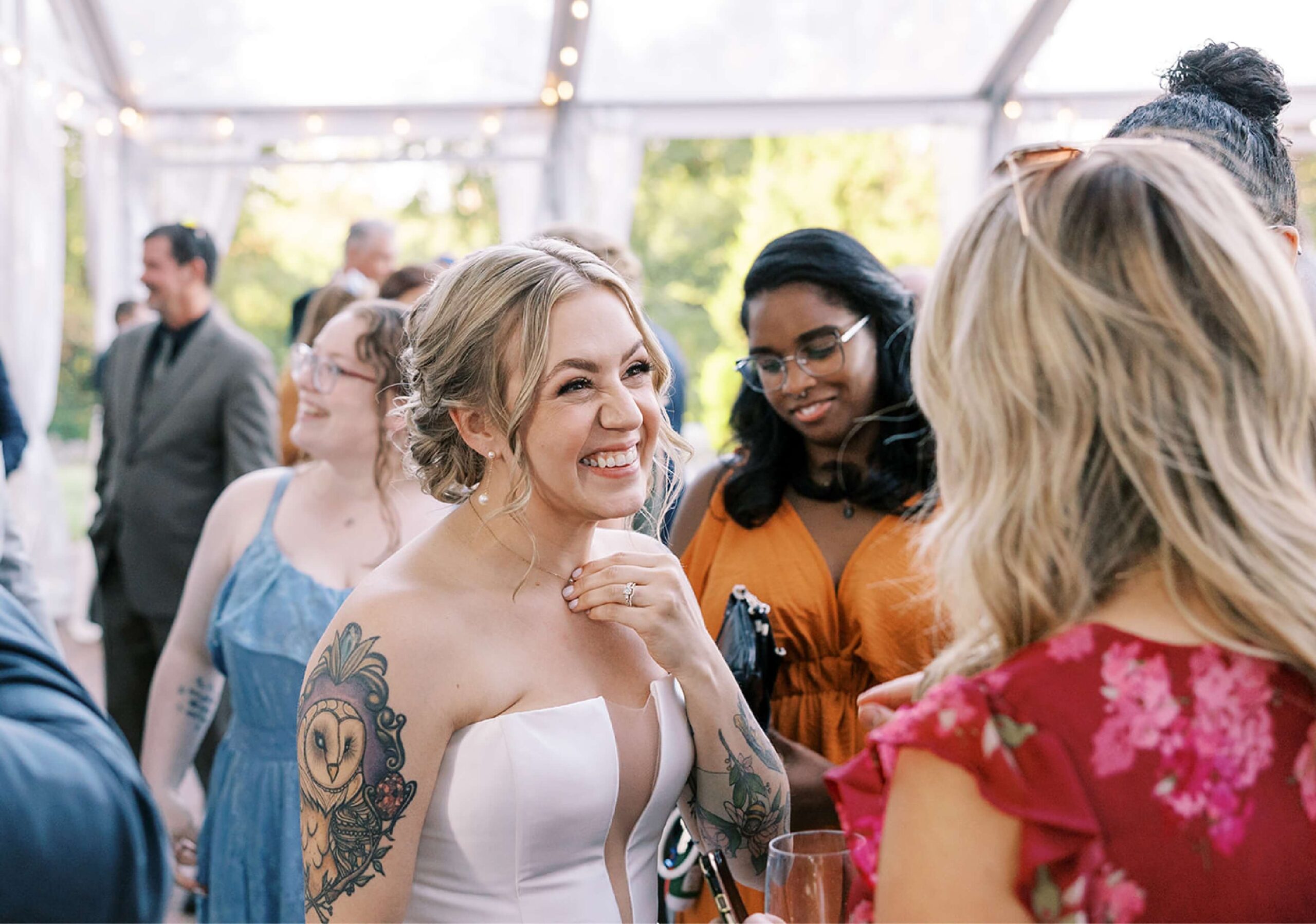 A smiling bride with tattoos converses with guests in a decorated venue, featuring string lights and greenery.